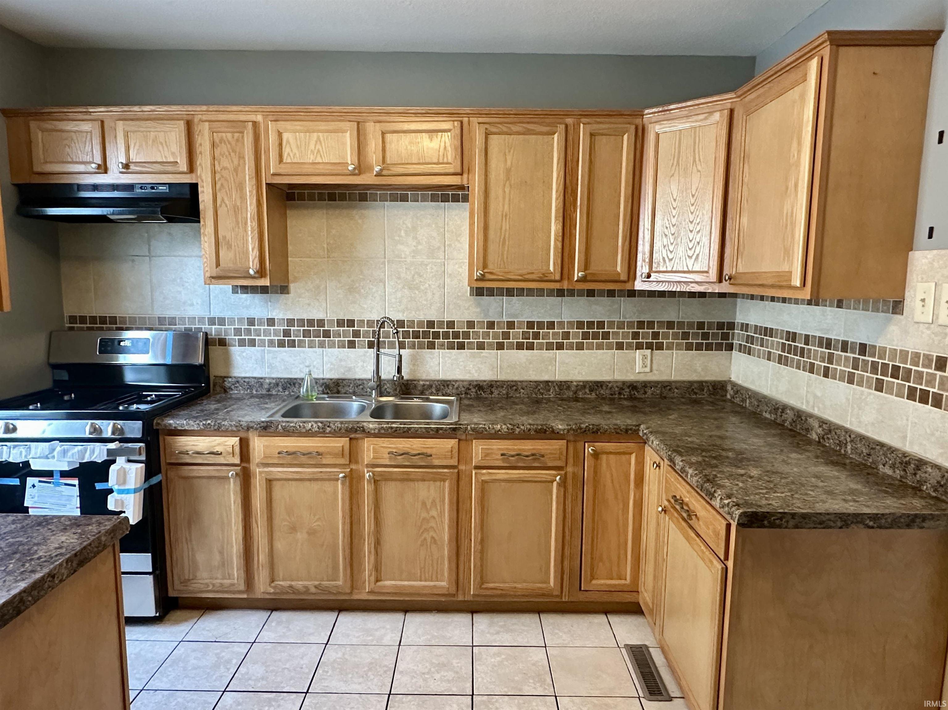 Kitchen featuring stainless steel range with gas stovetop, tasteful backsplash, dark countertops, under cabinet range hood, and light tile patterned floors