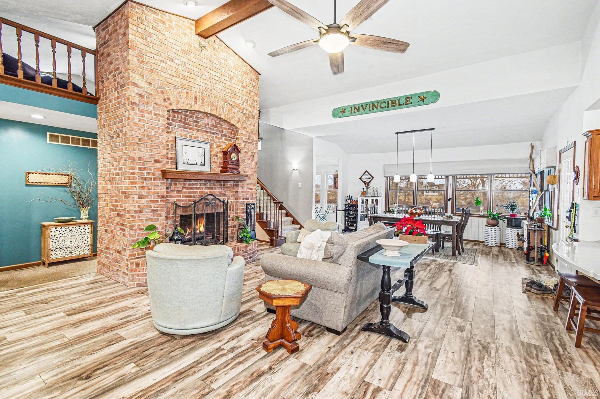Living room with a brick fireplace, beamed ceiling, light wood-style floors, high vaulted ceiling, and ceiling fan