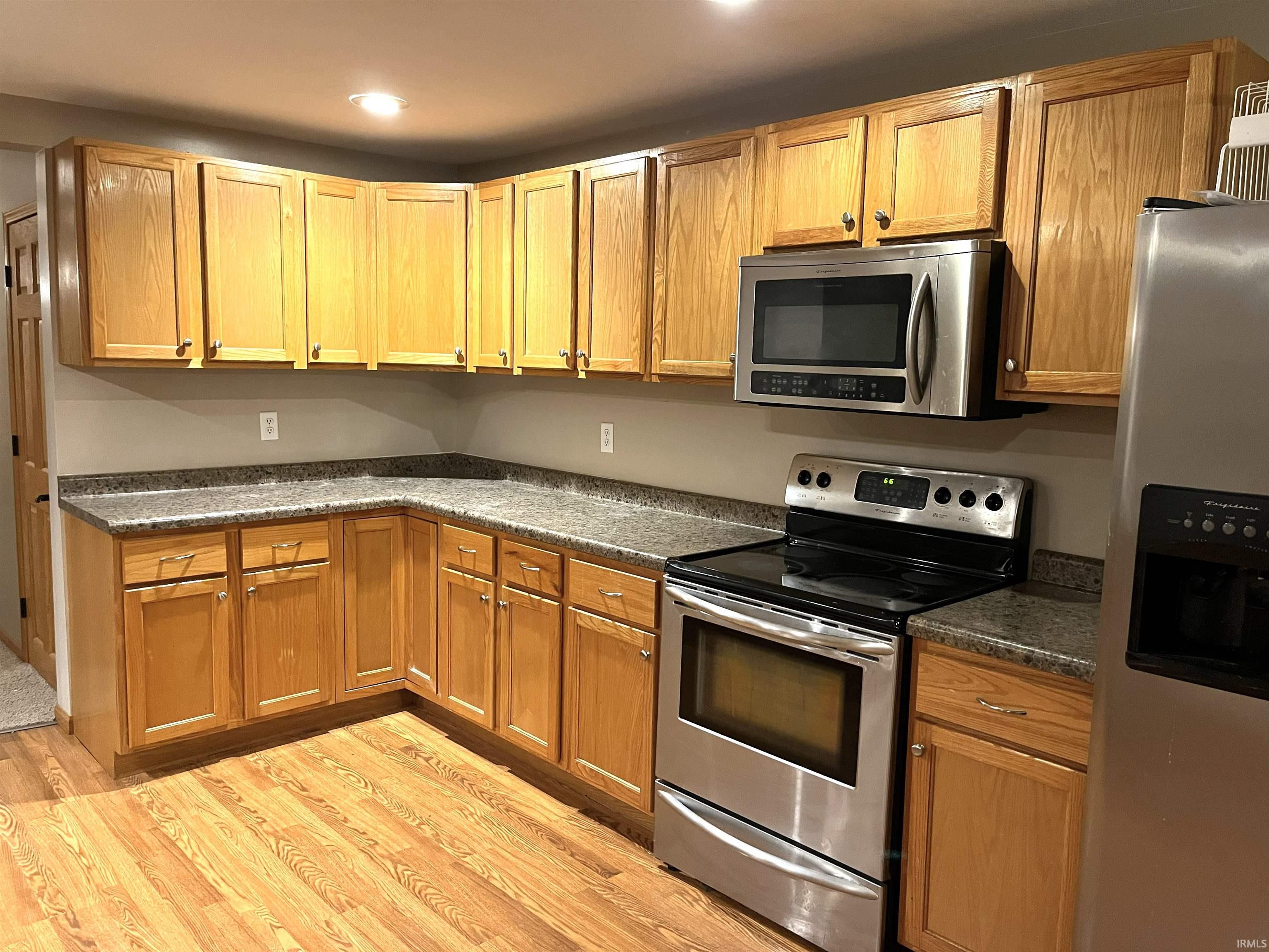 Kitchen with stainless steel appliances, light wood-style flooring, recessed lighting, brown cabinets, and dark stone countertops