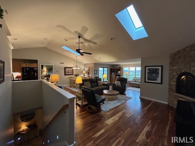 Living room with a skylight, vaulted ceiling, dark wood-style flooring, a ceiling fan, and a stone fireplace