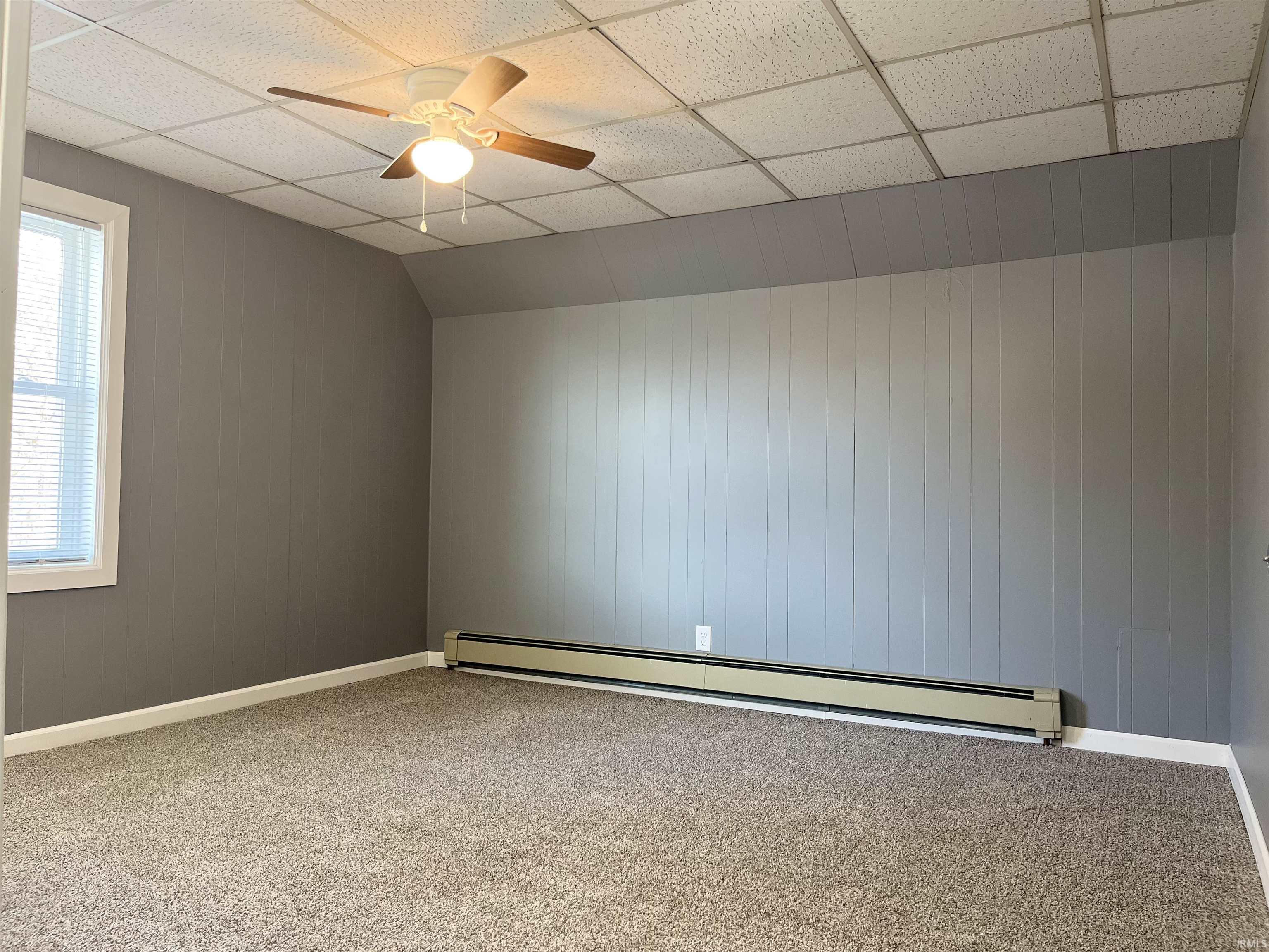 Carpeted empty room featuring wood walls, a baseboard radiator, a drop ceiling, ceiling fan, and lofted ceiling