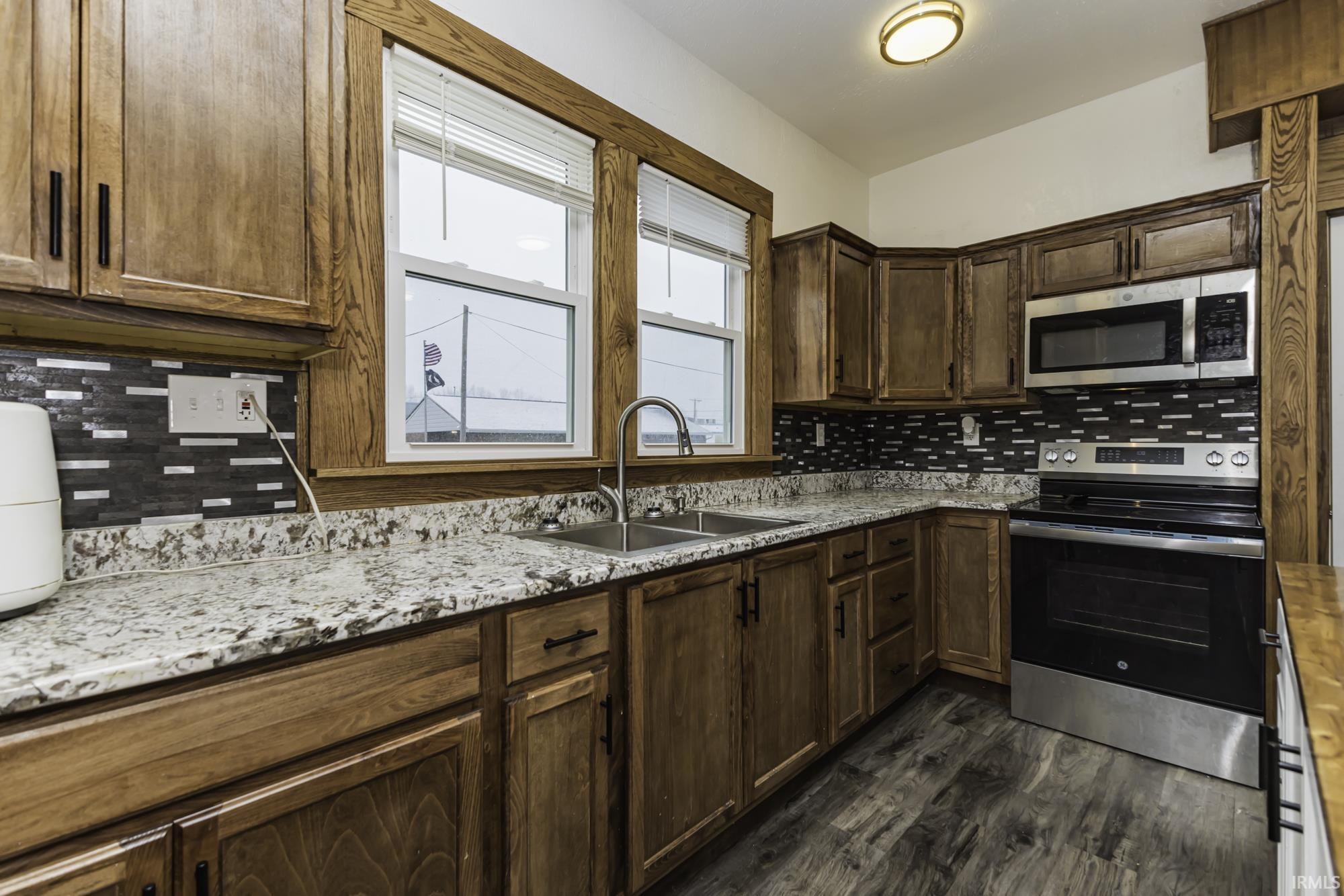 Kitchen featuring appliances with stainless steel finishes, backsplash, dark brown cabinetry, light stone counters, and dark wood finished floors