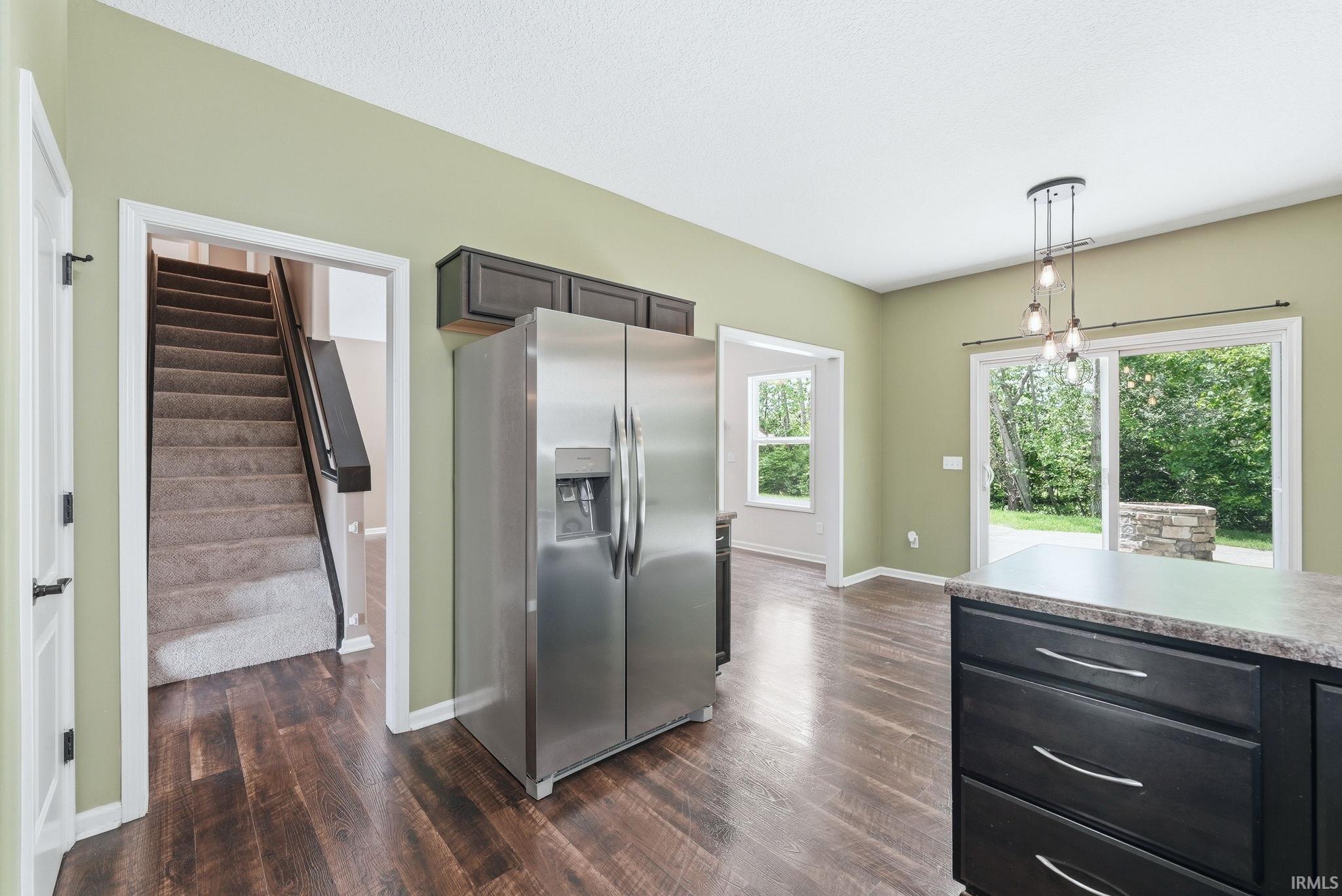 Kitchen space featuring wood-finish flooring, a stainless steel refrigerator, dark cabinetry with a light-toned countertop, and a contemporary multi-bulb pendant light fixture
