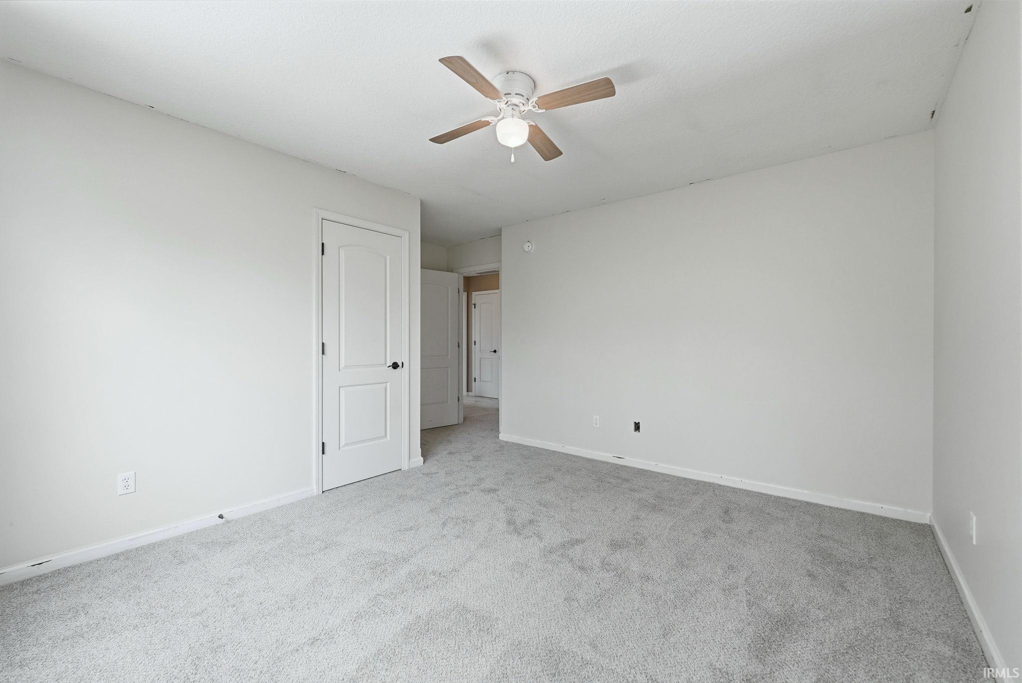 Spacious room featuring light gray carpeting, white walls, a ceiling fan with wood-finish blades, and a white paneled door with black hardware