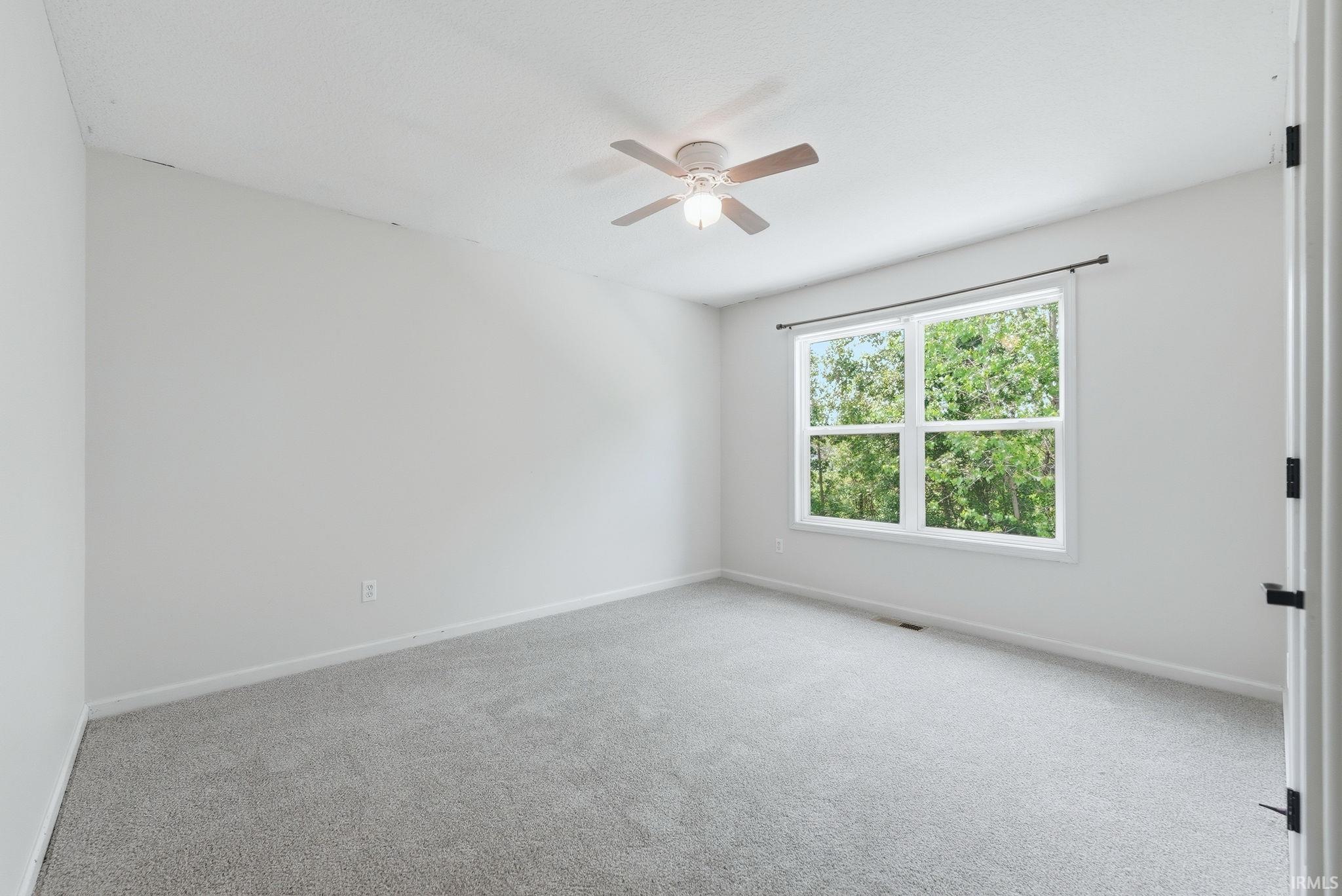 Spacious room featuring light gray carpeting, neutral wall tones, a large window with white trim, and a ceiling fan with light fixture