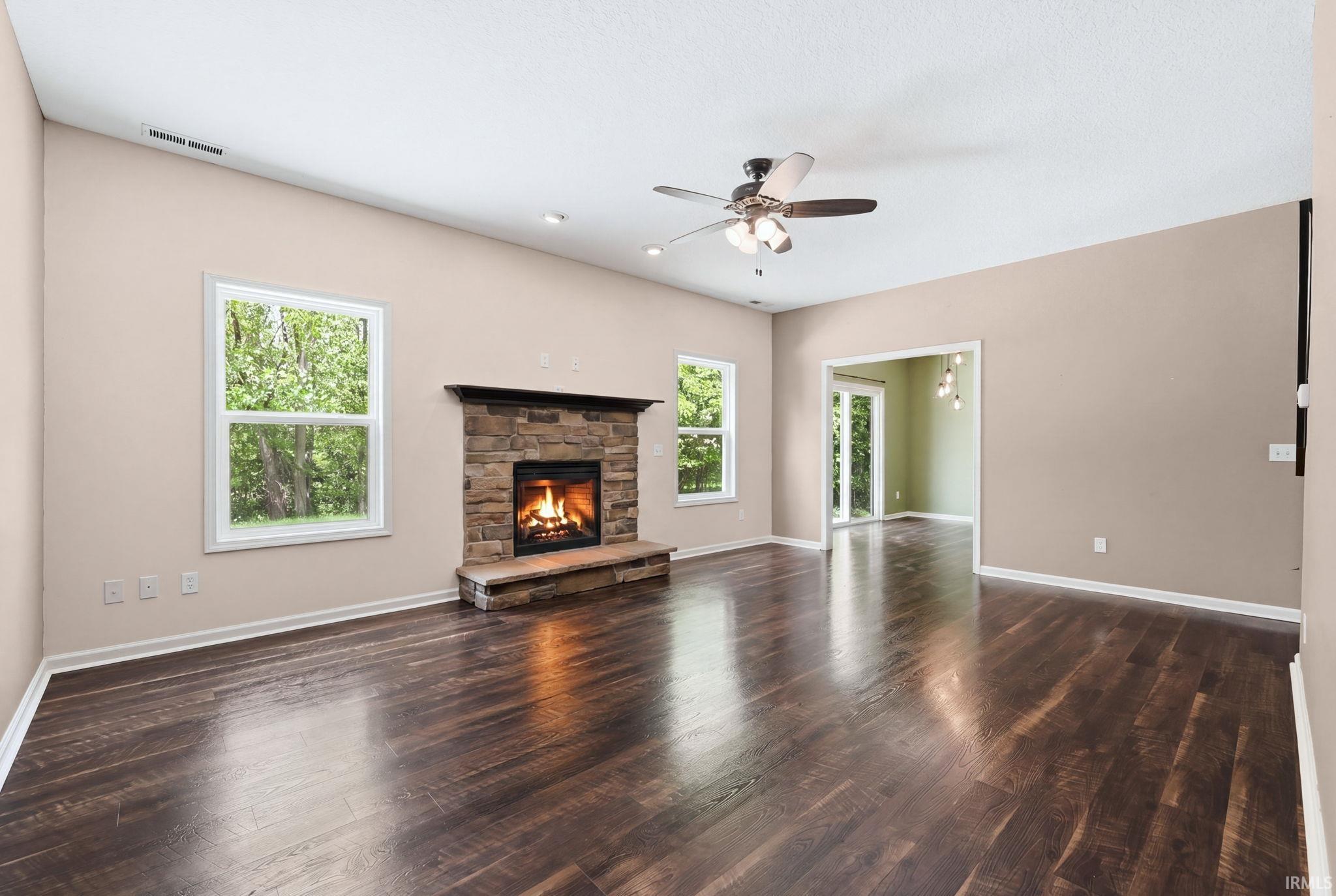 Spacious room featuring rich wood-finish flooring, a stone fireplace with a hearth, two large windows, a ceiling fan with integrated lighting, and recessed ceiling lights