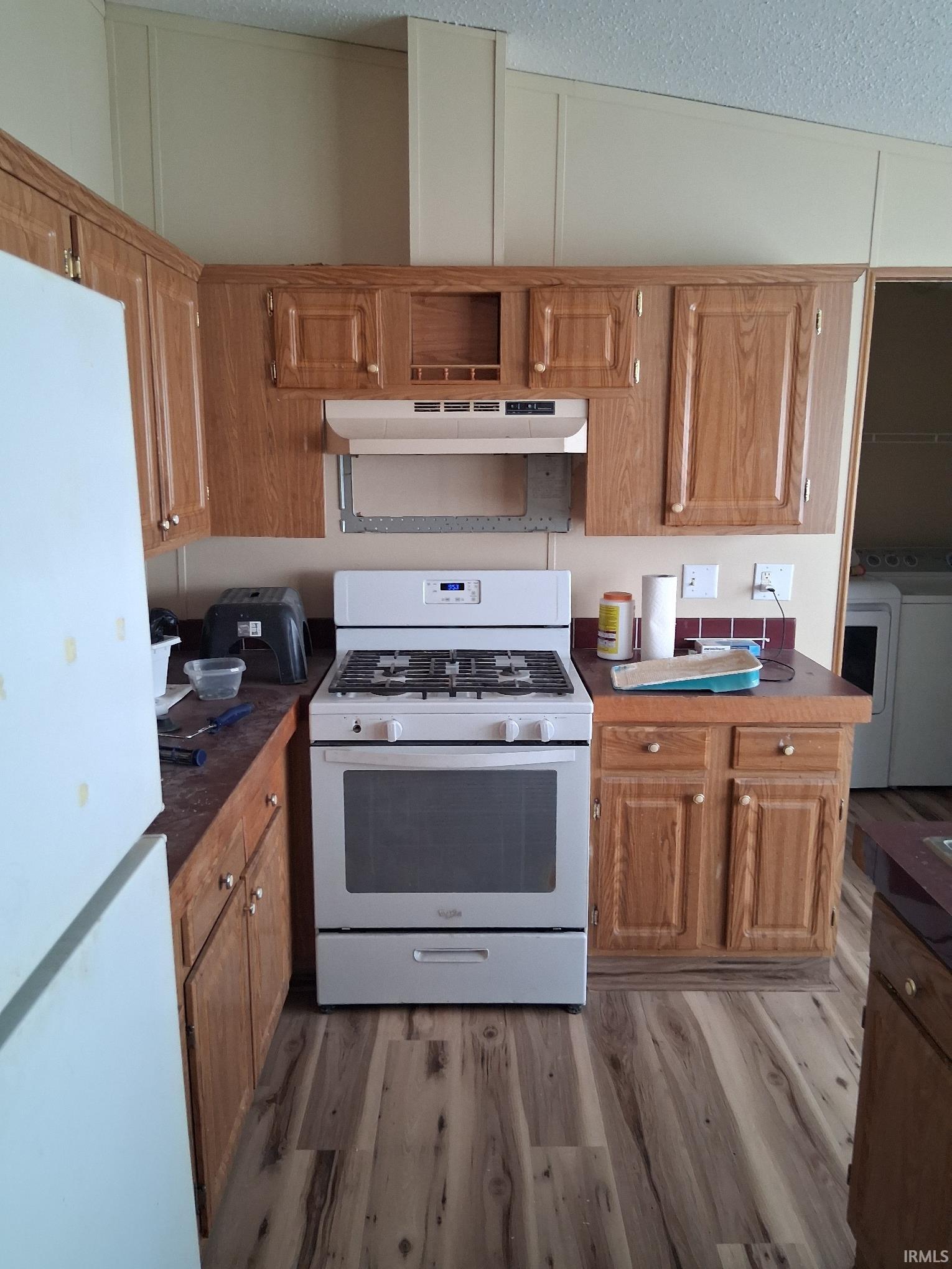 Kitchen with white appliances, brown cabinetry, under cabinet range hood, dark countertops, and a textured ceiling