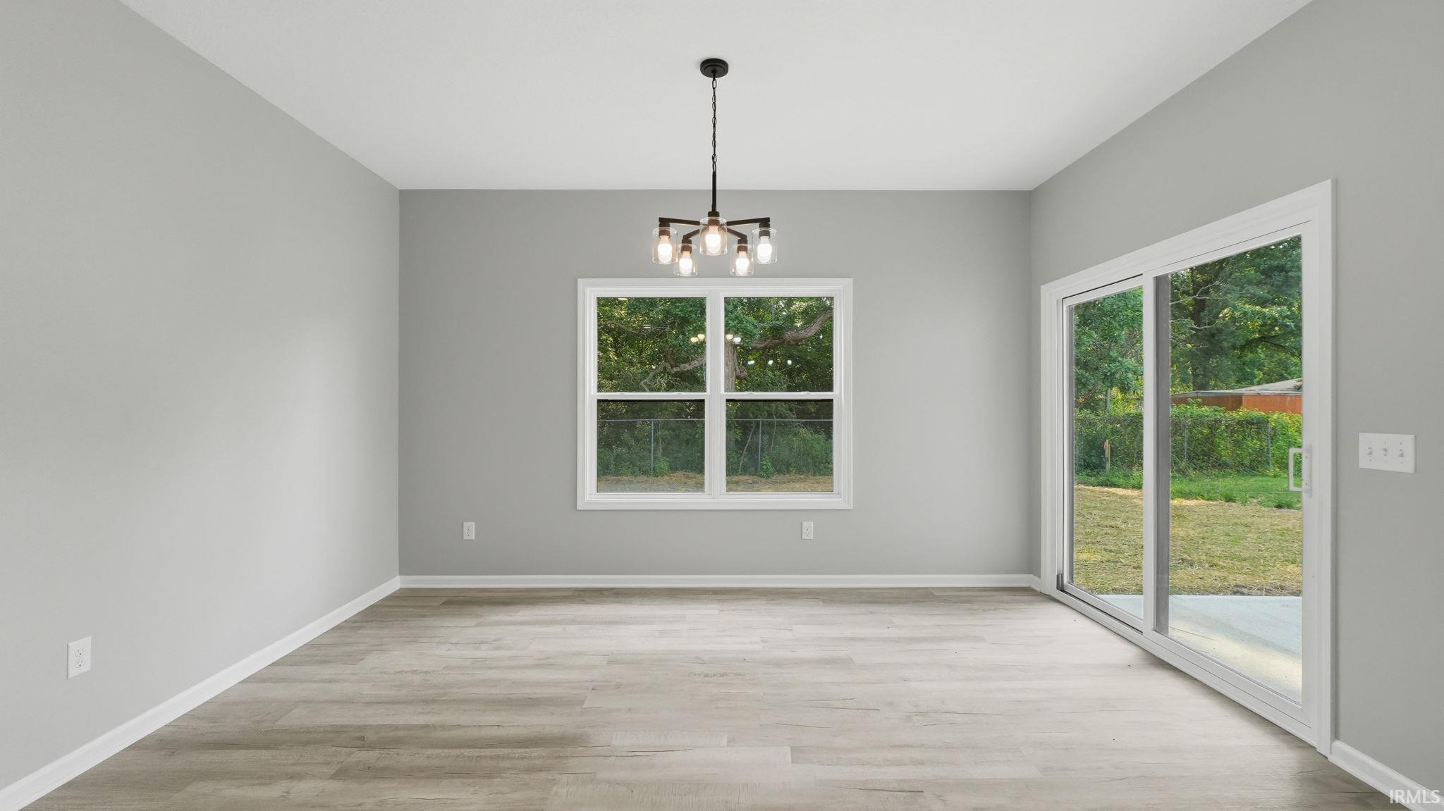 Spacious room featuring light gray walls, wood-finish flooring, a contemporary black chandelier, a large window, and a sliding glass door opening to outdoor space