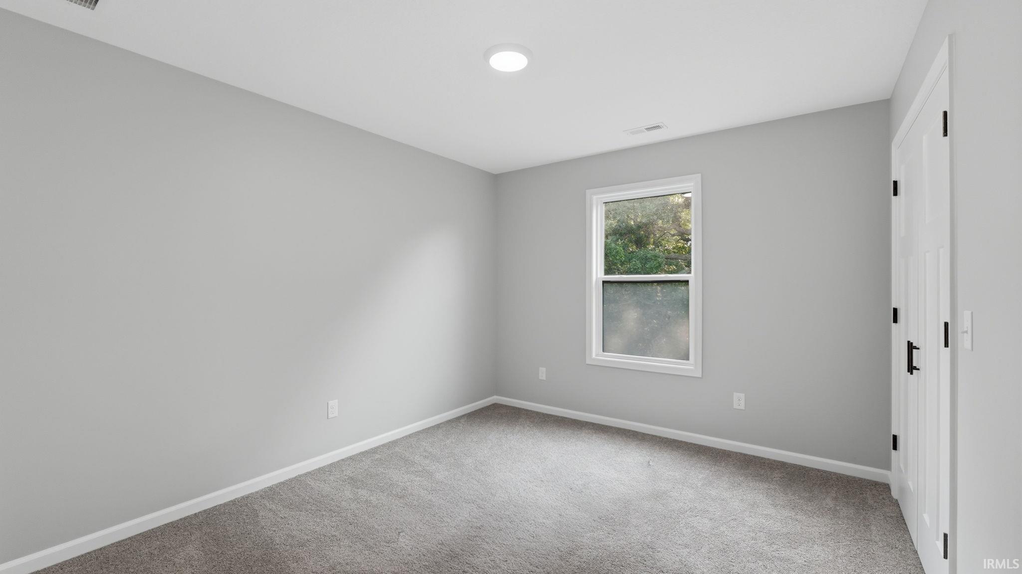 Spacious room with light gray walls, gray carpet, a single window, and a white door with black hardware