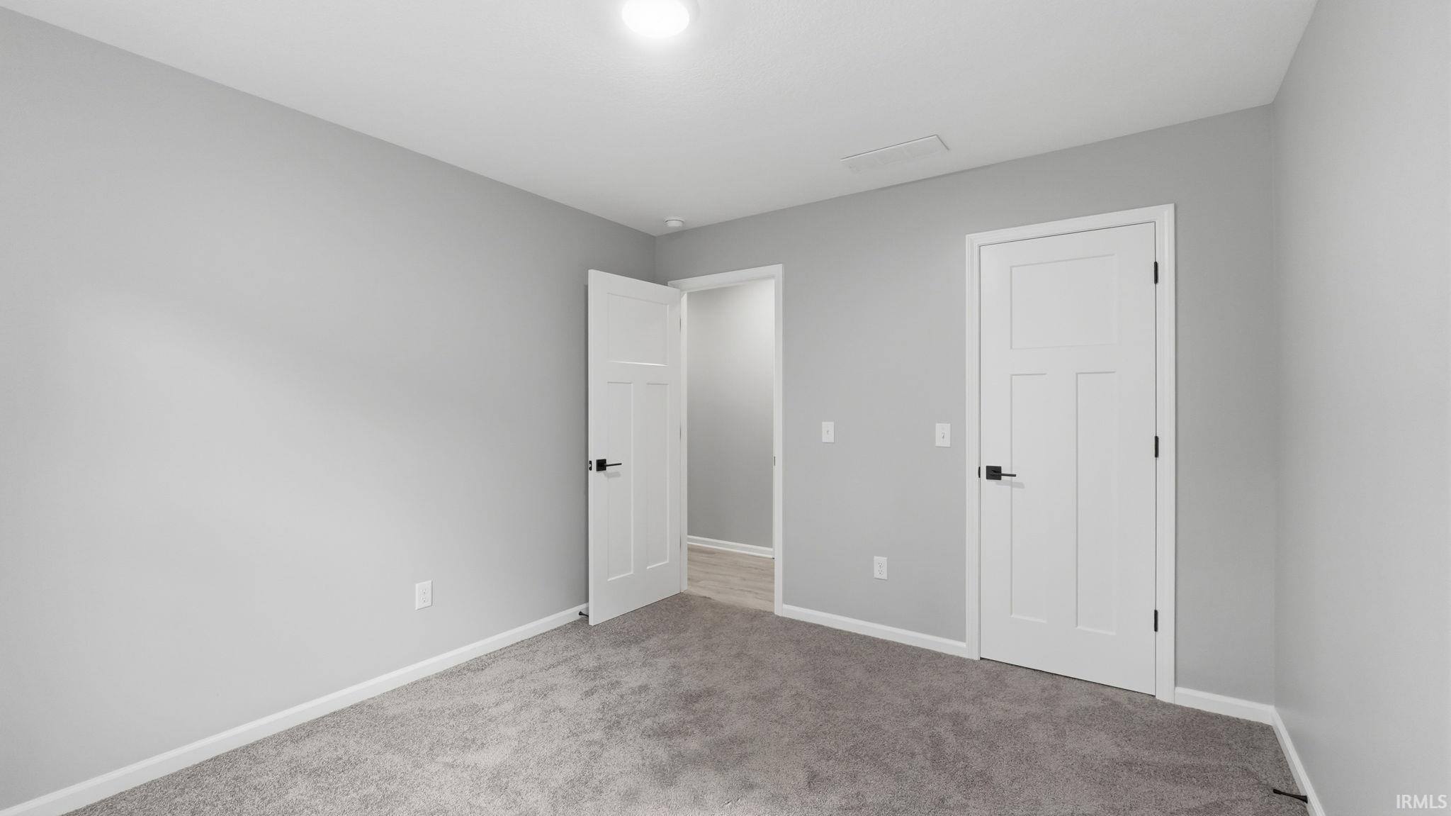 Spacious room featuring neutral gray walls, gray carpet flooring, white baseboards, and two contemporary white panel doors with black hardware