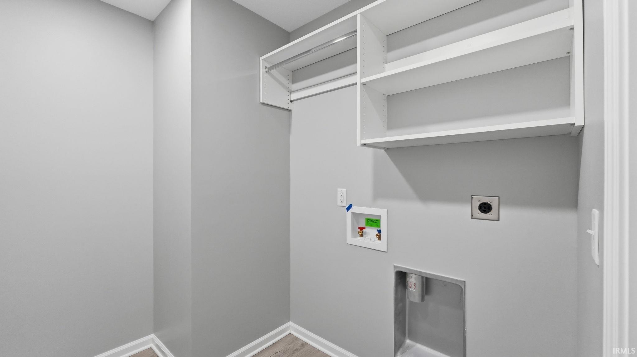 Dedicated laundry space featuring integrated shelving, a clothes hanging rod, and utility hookups, all complemented by light gray walls and wood-finish flooring