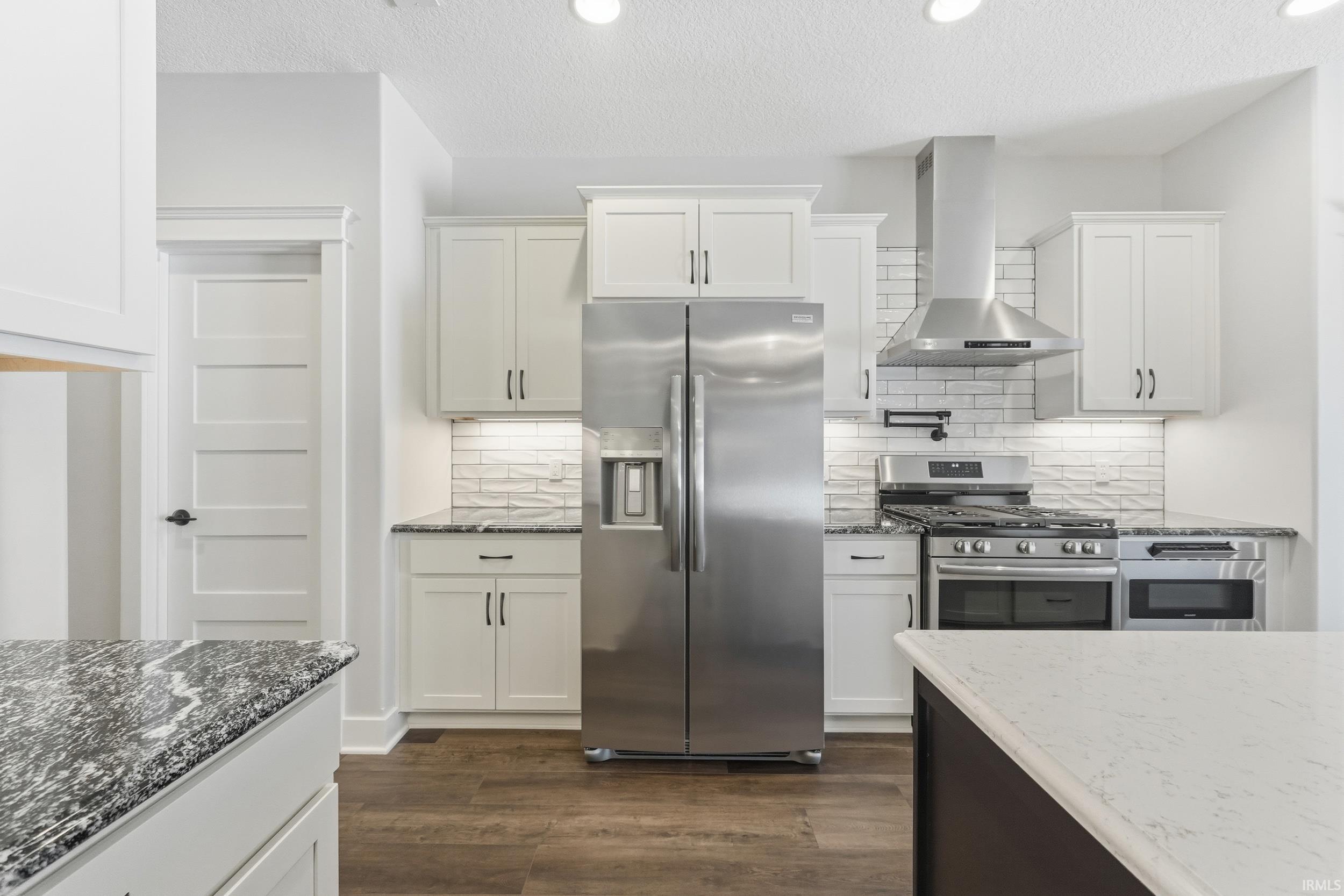 Kitchen featuring appliances with stainless steel finishes, dark stone countertops, wall chimney exhaust hood, a textured ceiling, and recessed lighting