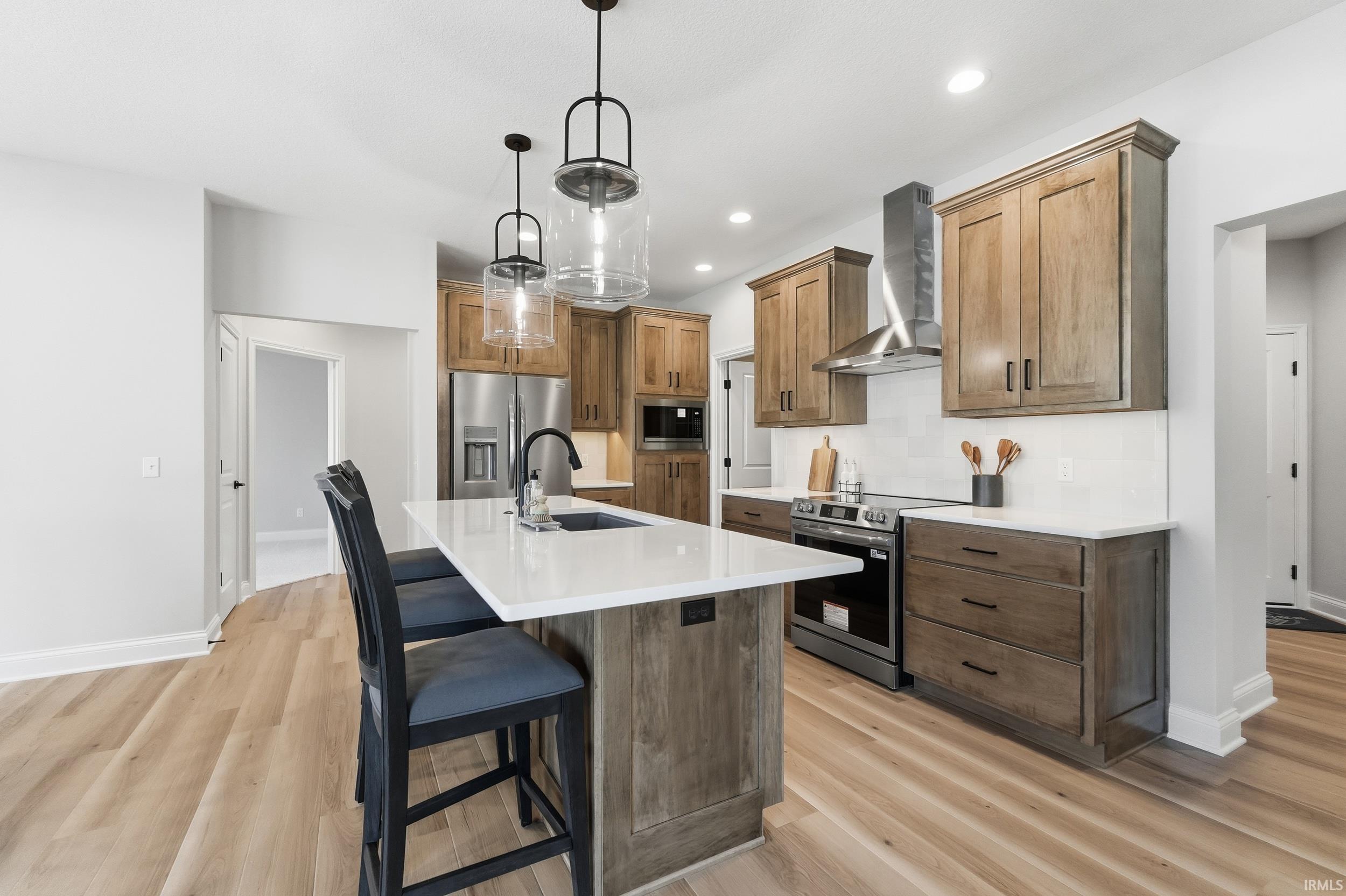 Kitchen featuring a kitchen bar, an island with sink, appliances with stainless steel finishes, pendant lighting, and wall chimney range hood