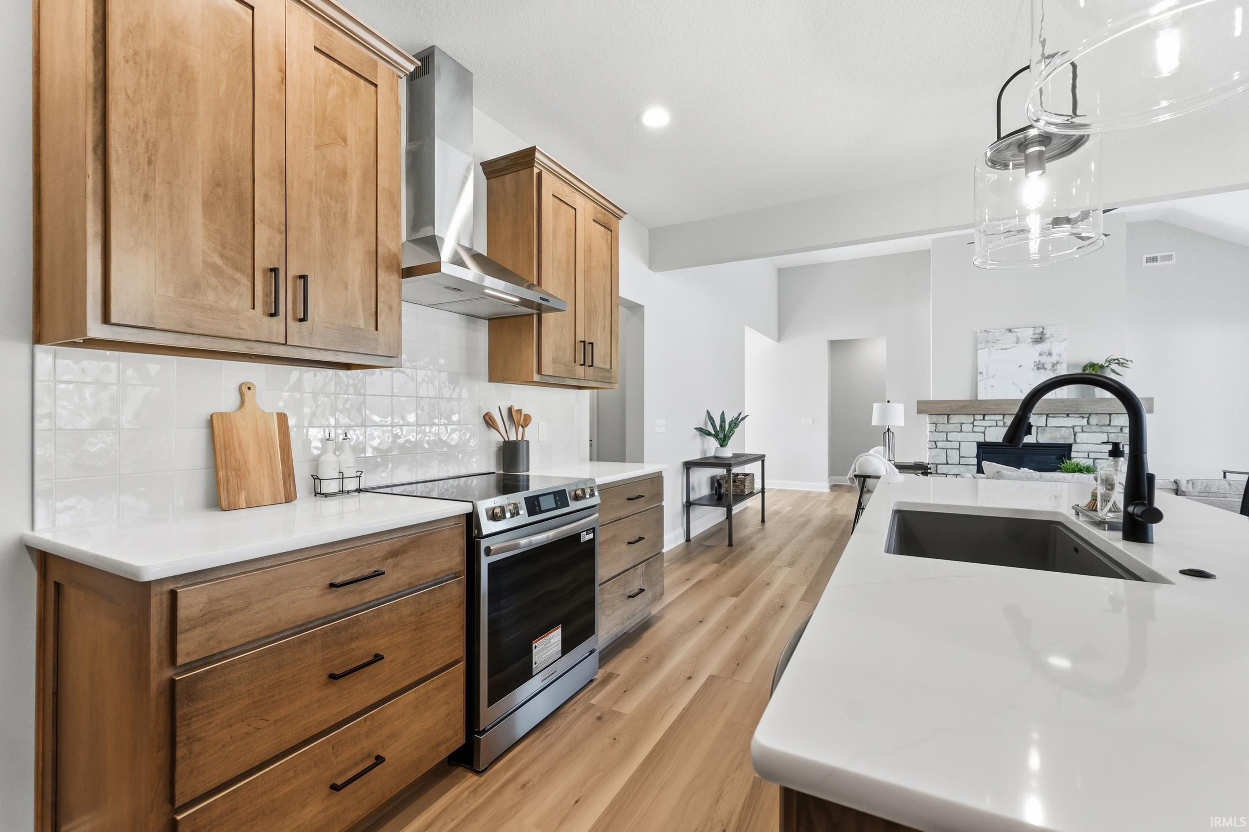 Kitchen with stainless steel range with electric stovetop, wall chimney exhaust hood, light wood finished floors, light stone counters, and hanging light fixtures