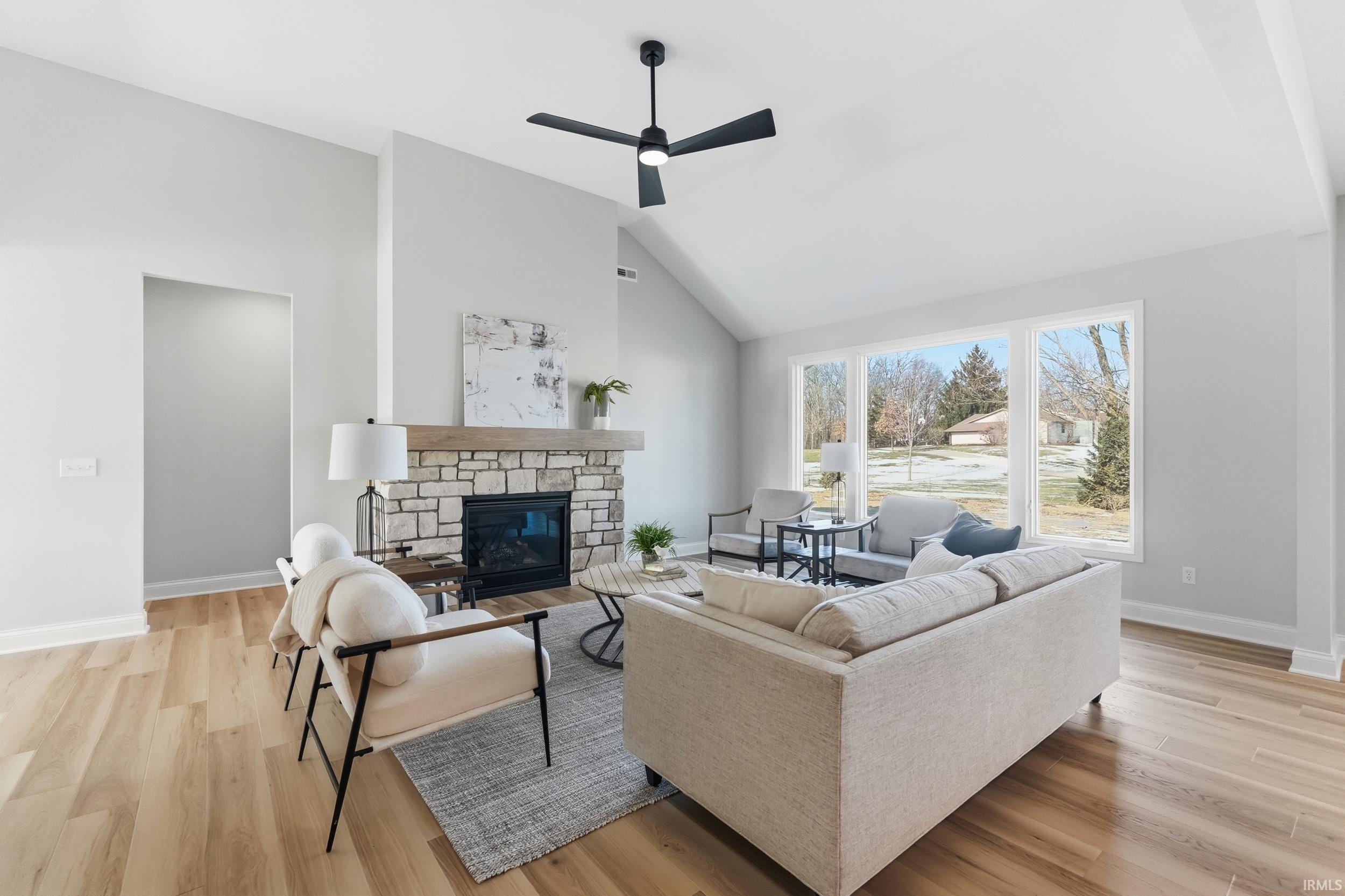 Living area with a stone fireplace, light wood finished floors, high vaulted ceiling, and a ceiling fan