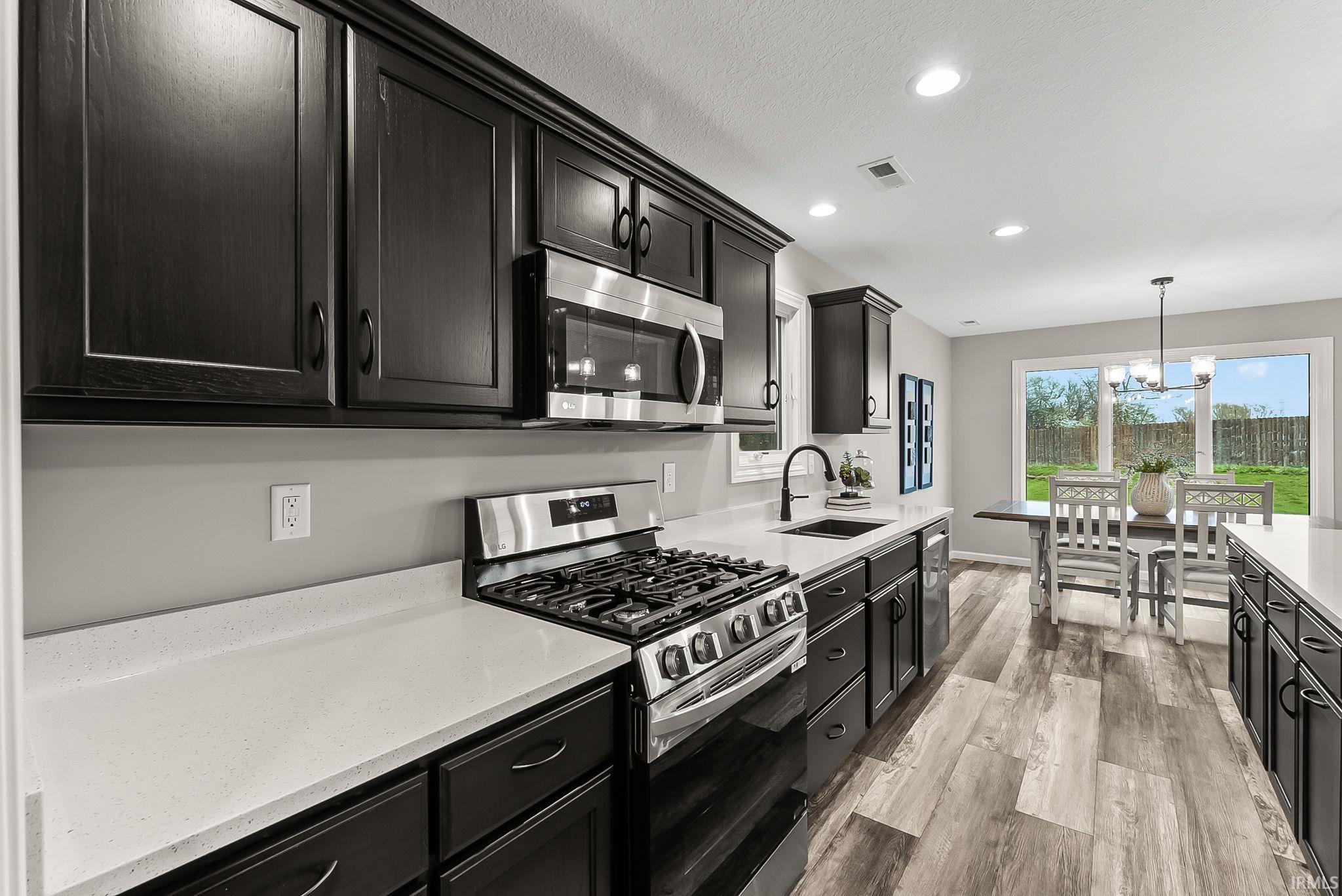 Kitchen with dark cabinets, stainless steel appliances, light wood-style flooring, light stone counters, and a chandelier