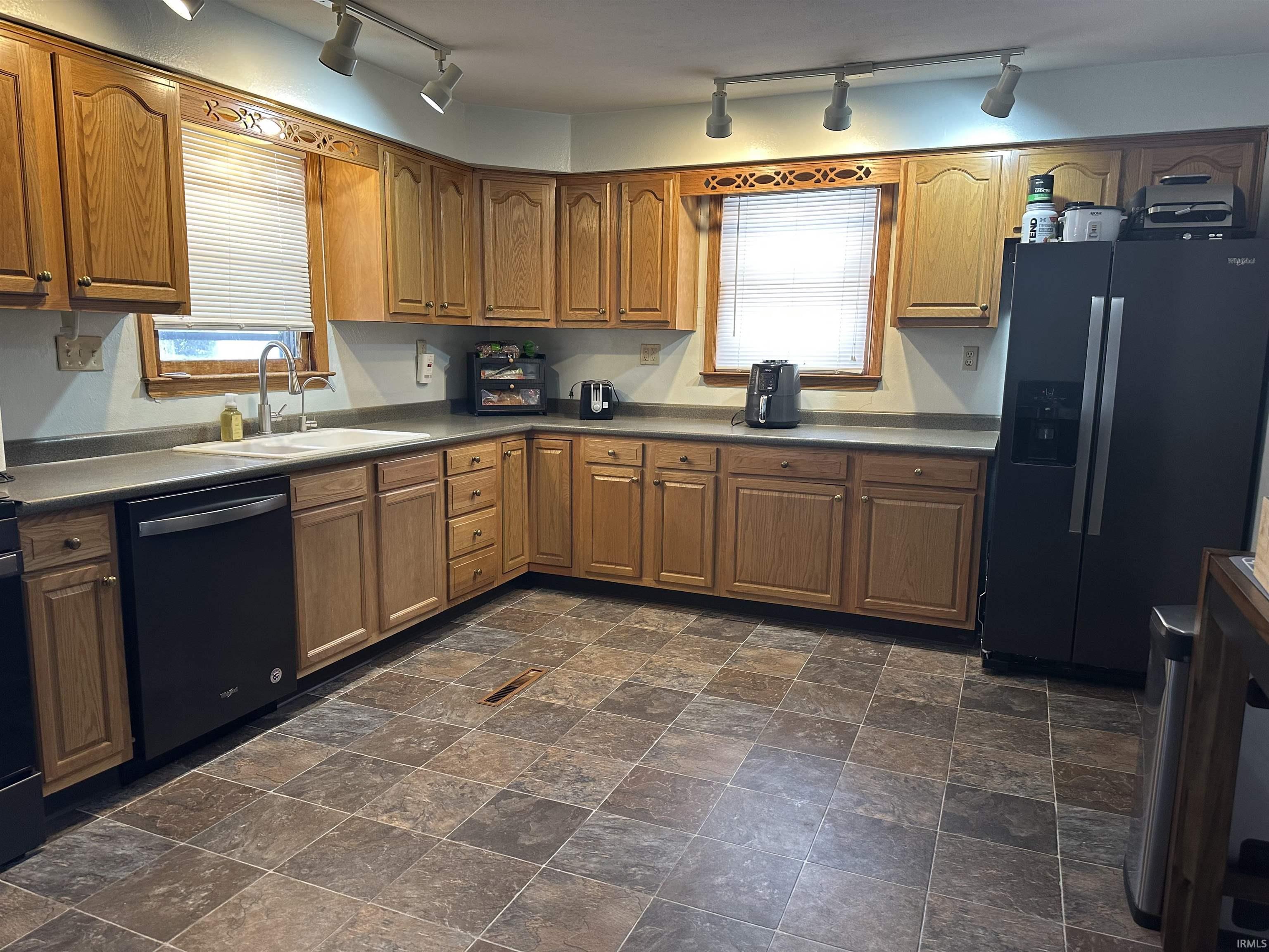 Kitchen featuring black fridge, dishwasher, stone finish flooring, brown cabinetry, and plenty of natural light
