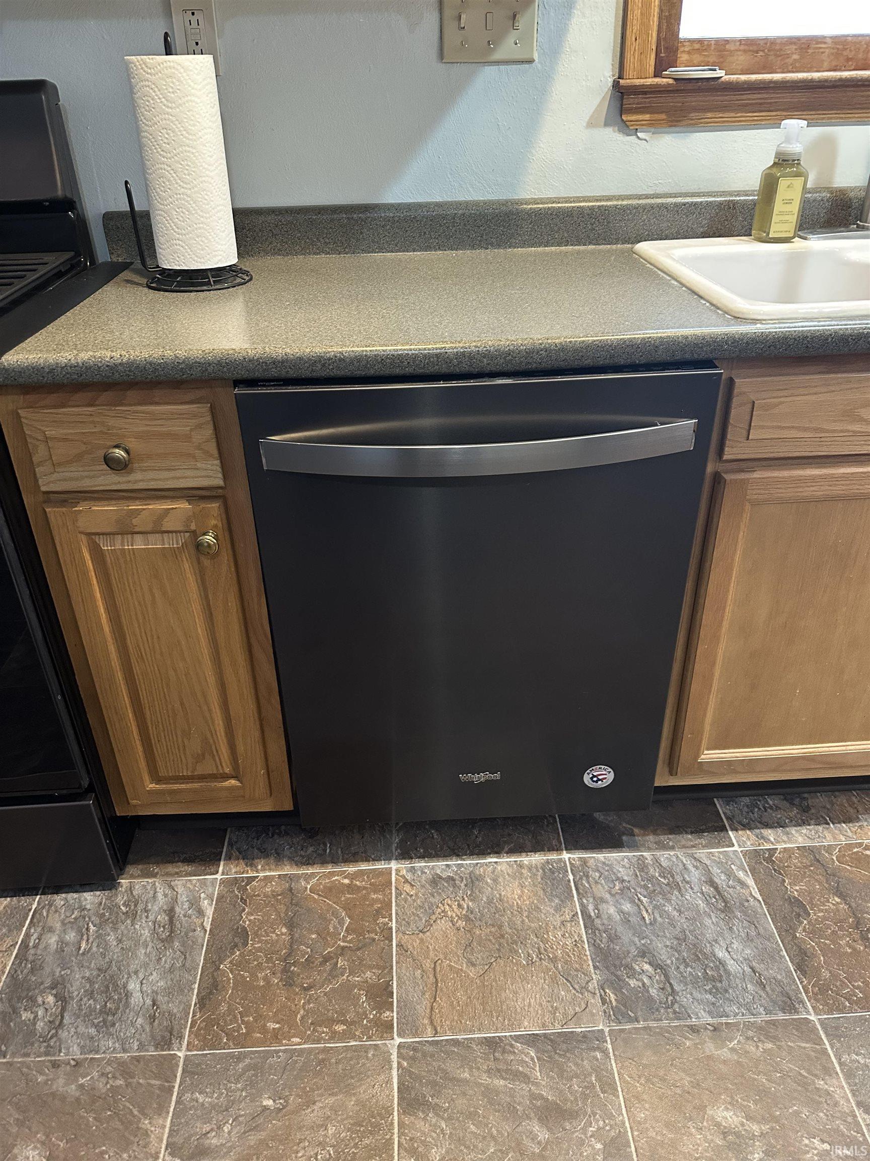 Kitchen view of stainless steel dishwasher, brown cabinetry, dark countertops, and black range oven