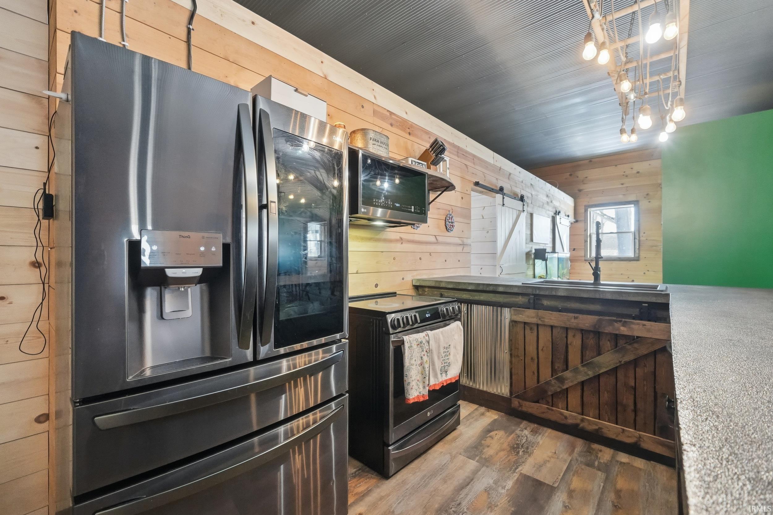 Kitchen featuring wood walls, a barn door, stainless steel appliances, dark wood finished floors, and hanging light fixtures