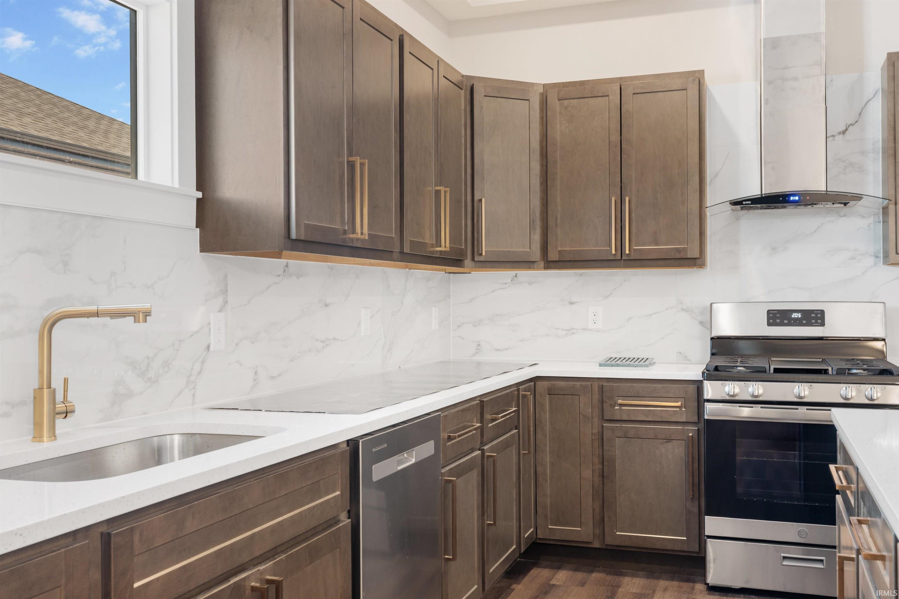 Kitchen with appliances with stainless steel finishes, dark brown cabinetry, wall chimney exhaust hood, light stone countertops, and backsplash