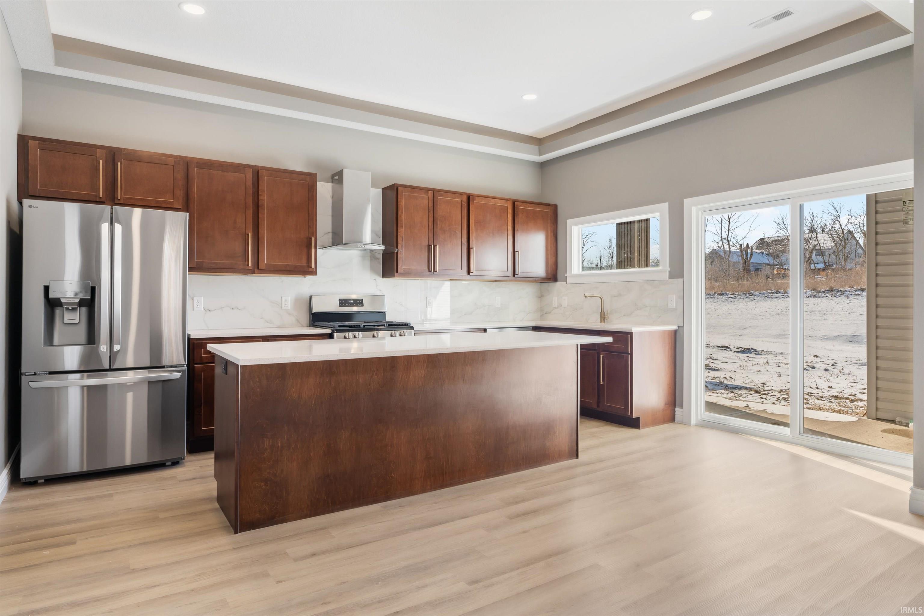 Kitchen with appliances with stainless steel finishes, a center island, wall chimney exhaust hood, tasteful backsplash, and light wood-style floors