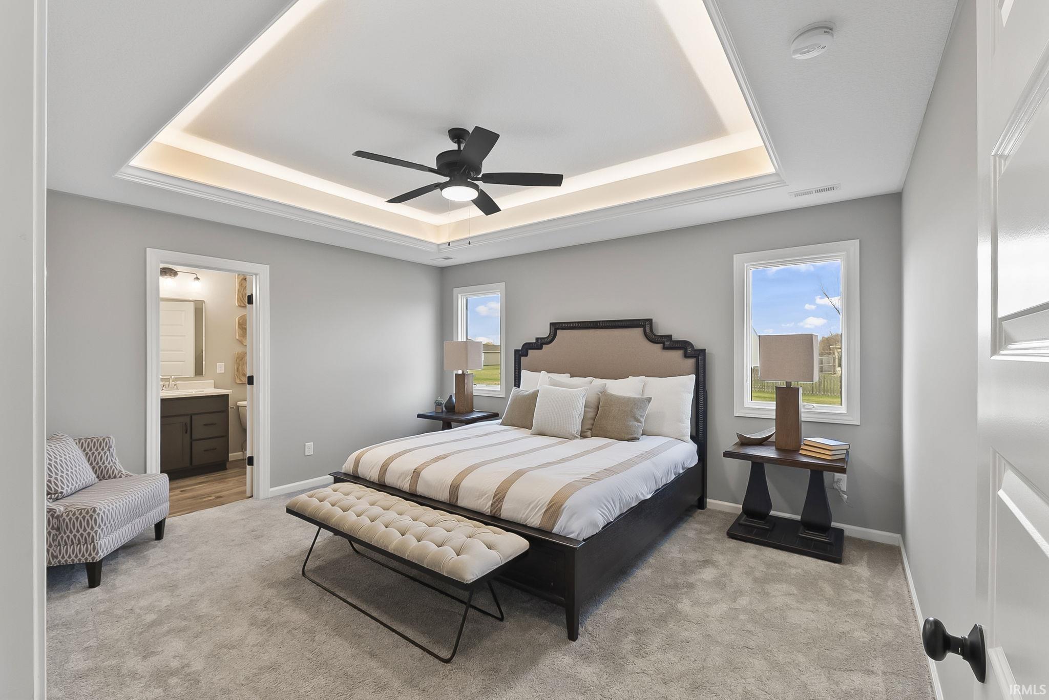 Bedroom with light colored carpet, multiple windows, a ceiling fan, ensuite bath, and a tray ceiling