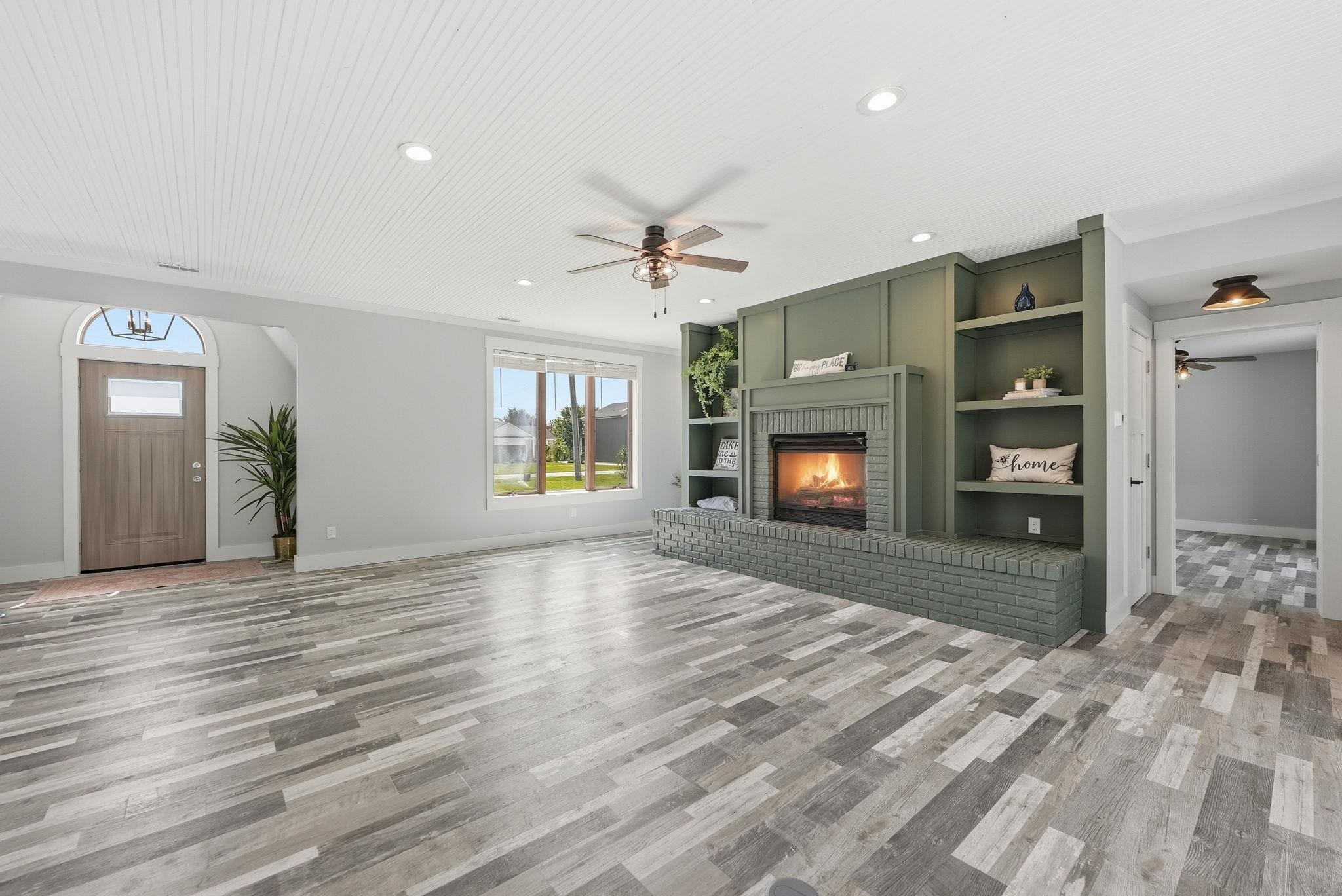 Spacious living area featuring plank-style flooring, a decorative fireplace with built-in shelving, large windows providing natural light, and a beadboard ceiling with recessed lighting and ceiling fans