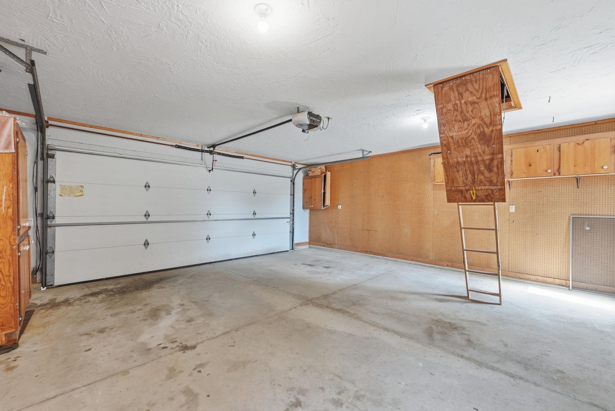 Garage interior featuring a sectional white garage door with opener, concrete flooring, wood-paneled walls, built-in upper wood cabinetry, and a pull-down attic access ladder
