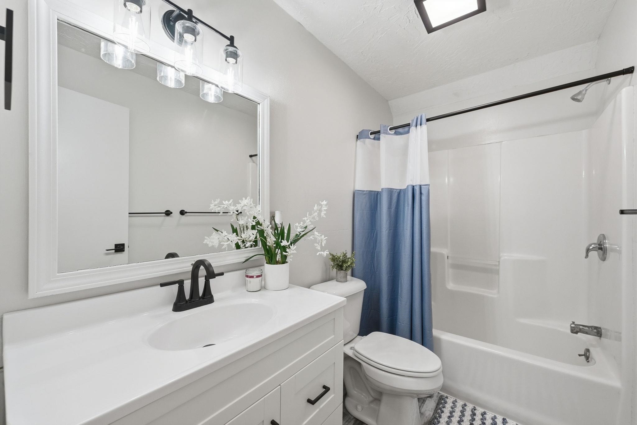 Bathroom featuring a white vanity with an integrated sink, a rectangular framed mirror, matte black fixtures, a three-light vanity bar, and a white bathtub with a shower
