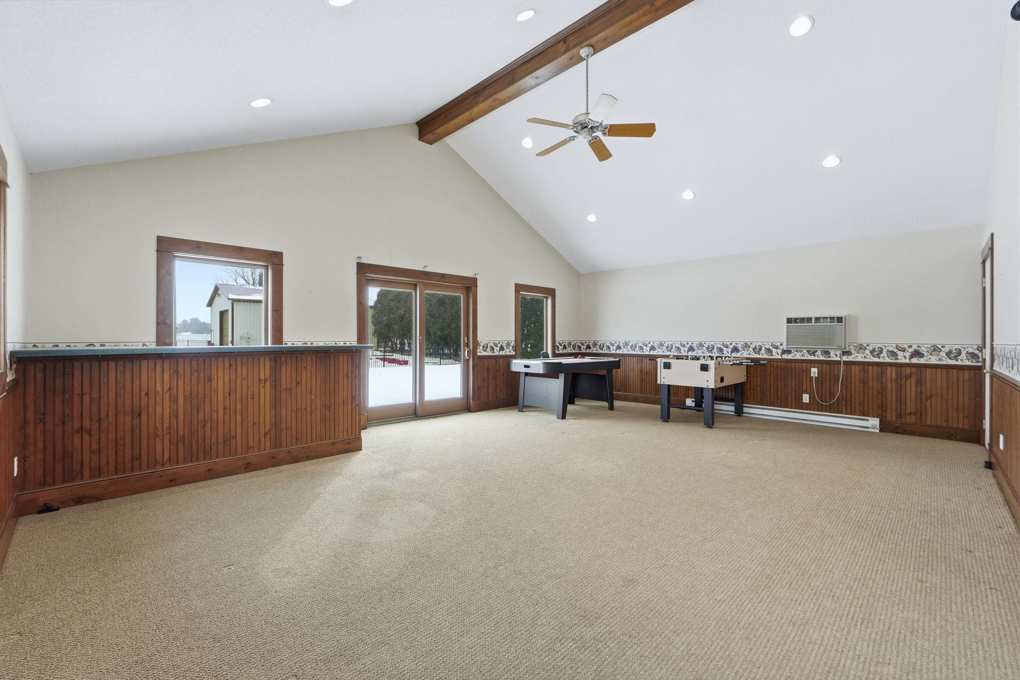 Spacious vaulted room featuring exposed wood beams, recessed lighting, a ceiling fan, wood-paneled wainscoting, and glass sliding doors leading to the exterior