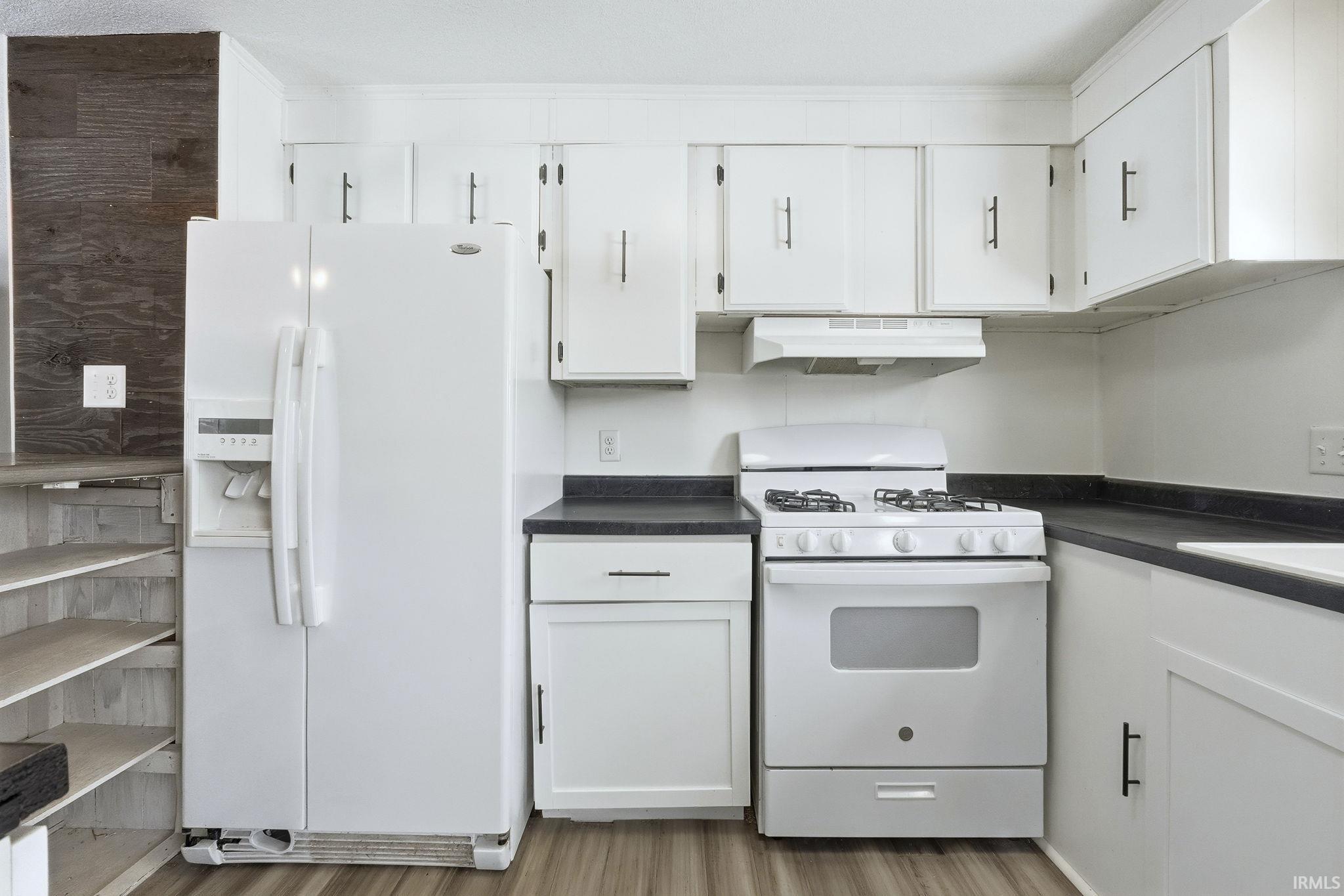 Kitchen with dark countertops, white appliances, white cabinetry, under cabinet range hood, and light wood-style flooring