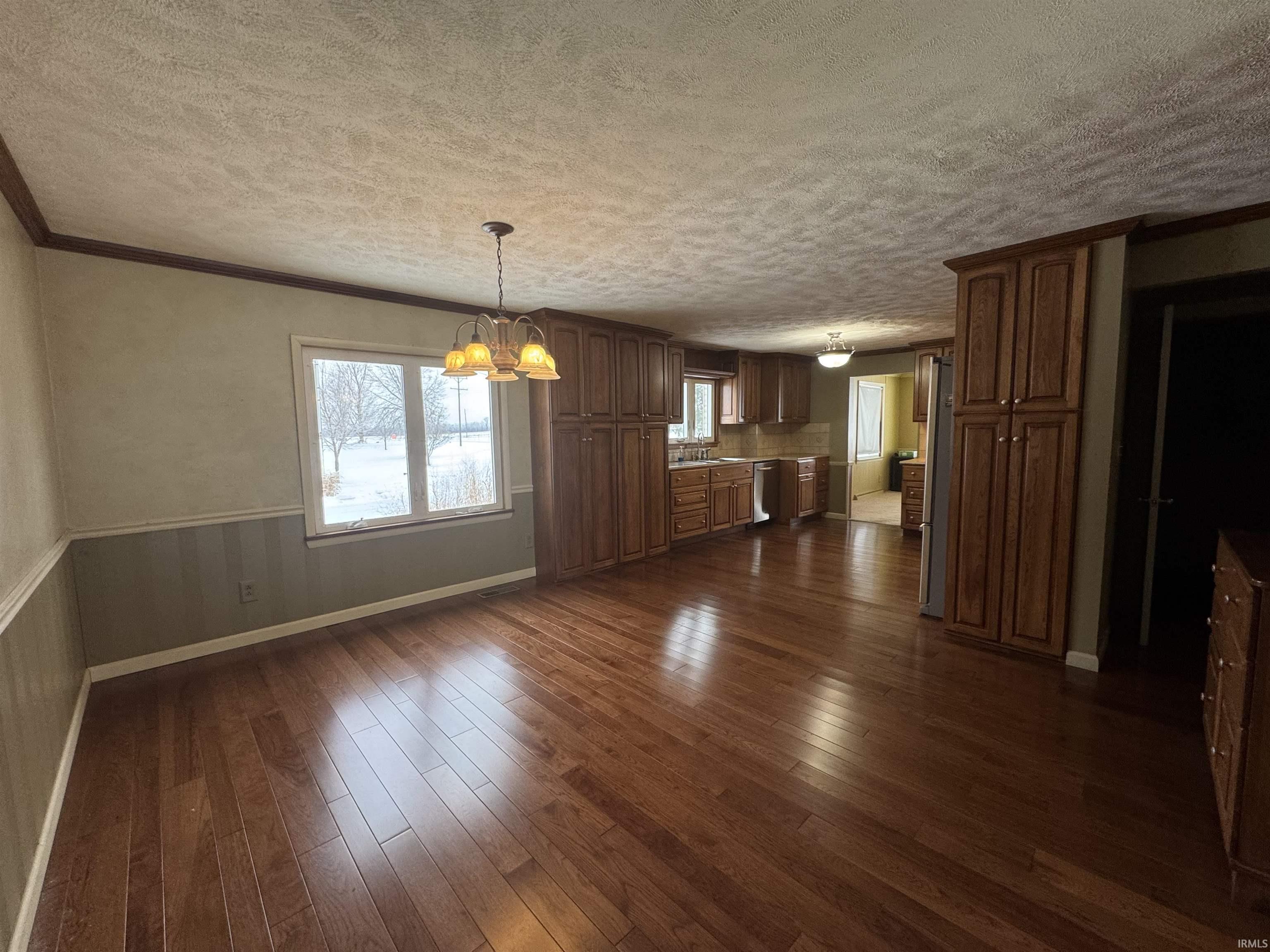 Unfurnished dining area featuring wainscoting, a chandelier, dark wood-type flooring, ornamental molding, and a textured ceiling