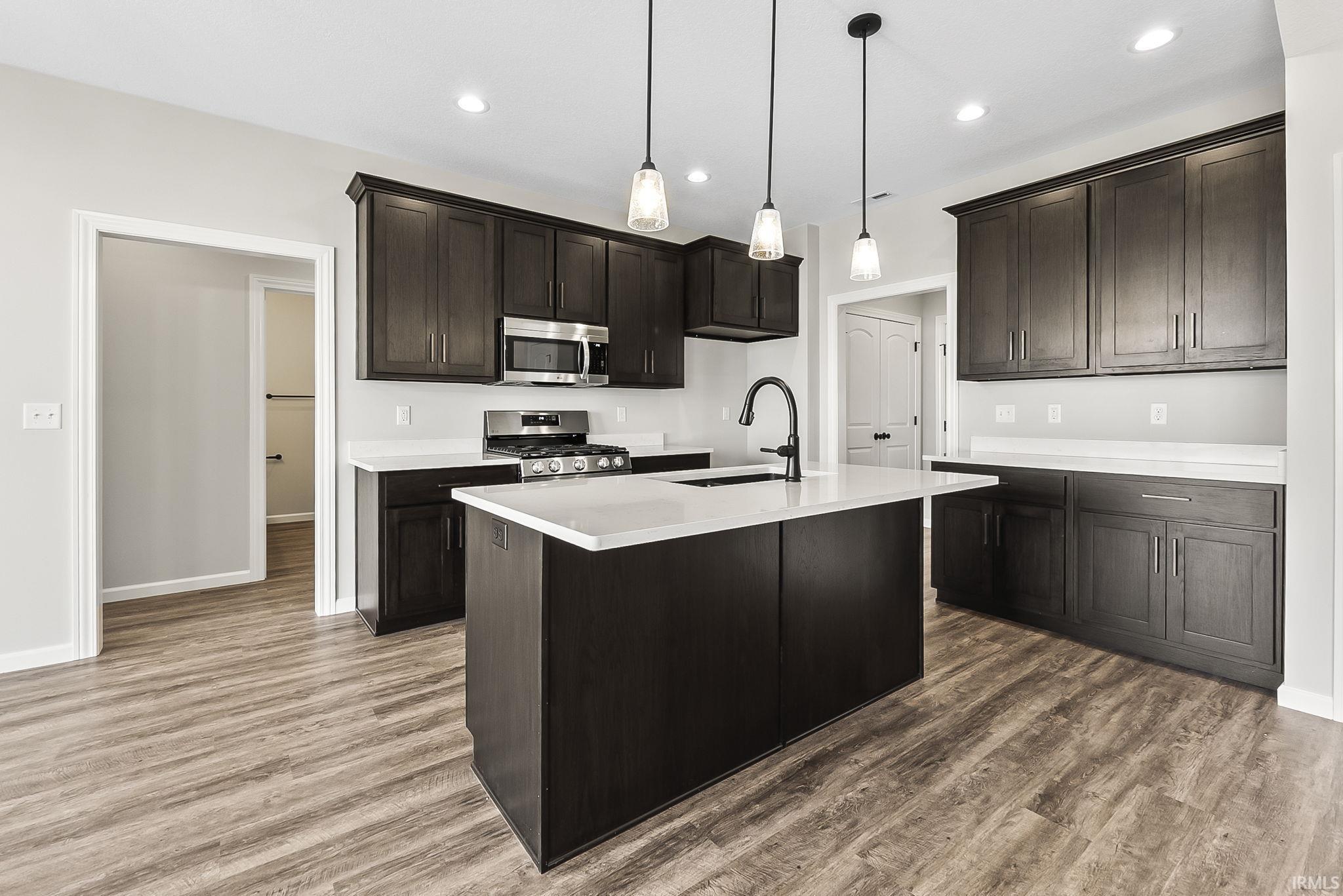Kitchen featuring stainless steel appliances, an island with sink, dark wood-type flooring, dark wood finish cabinetry, and decorative light fixtures