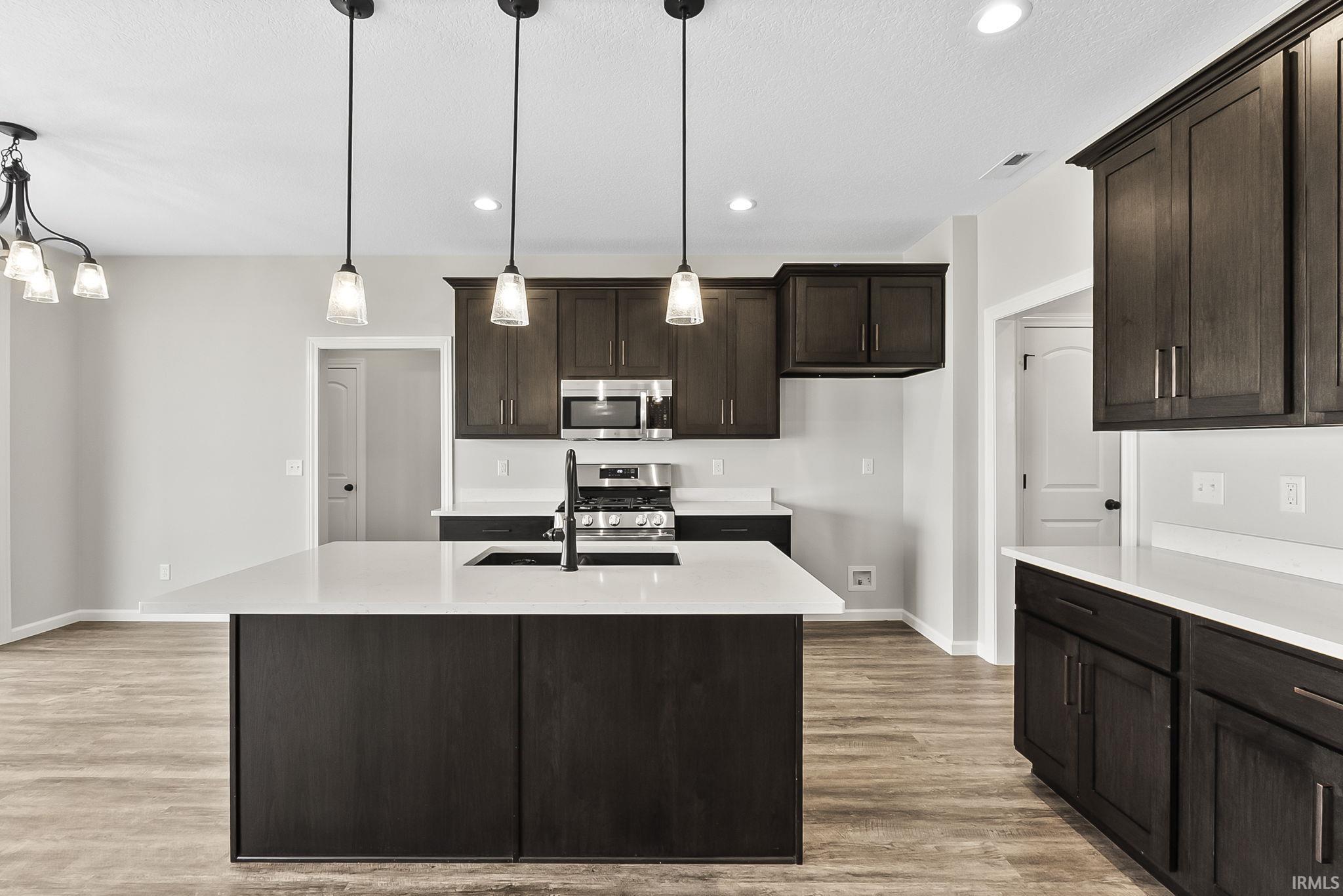 Kitchen with dark wood finish cabinets, stainless steel appliances, light wood-style floors, light stone countertops, and a center island with sink