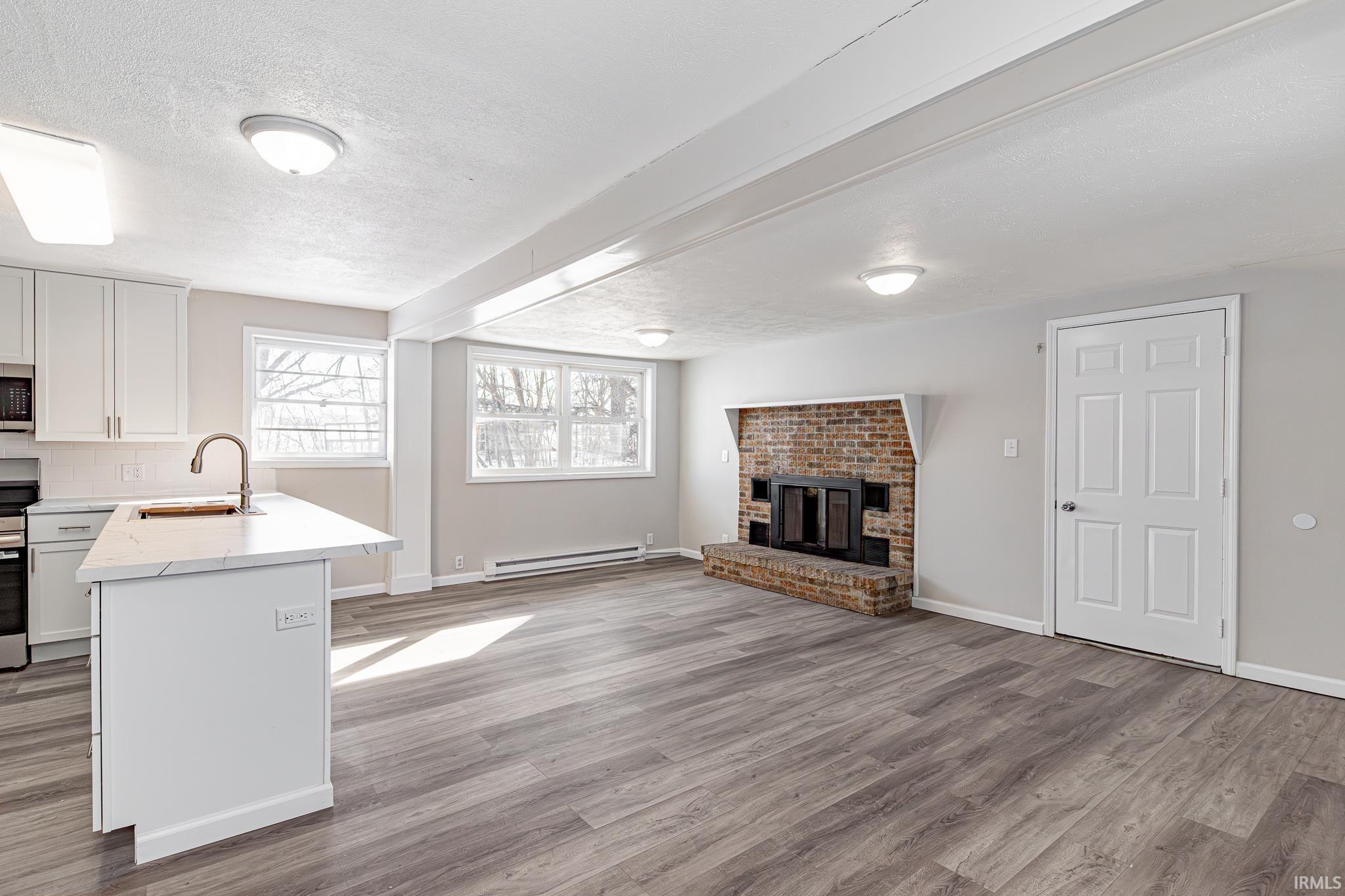 Unfurnished living room with a textured ceiling, light wood finished floors, a baseboard radiator, a brick fireplace, and beam ceiling