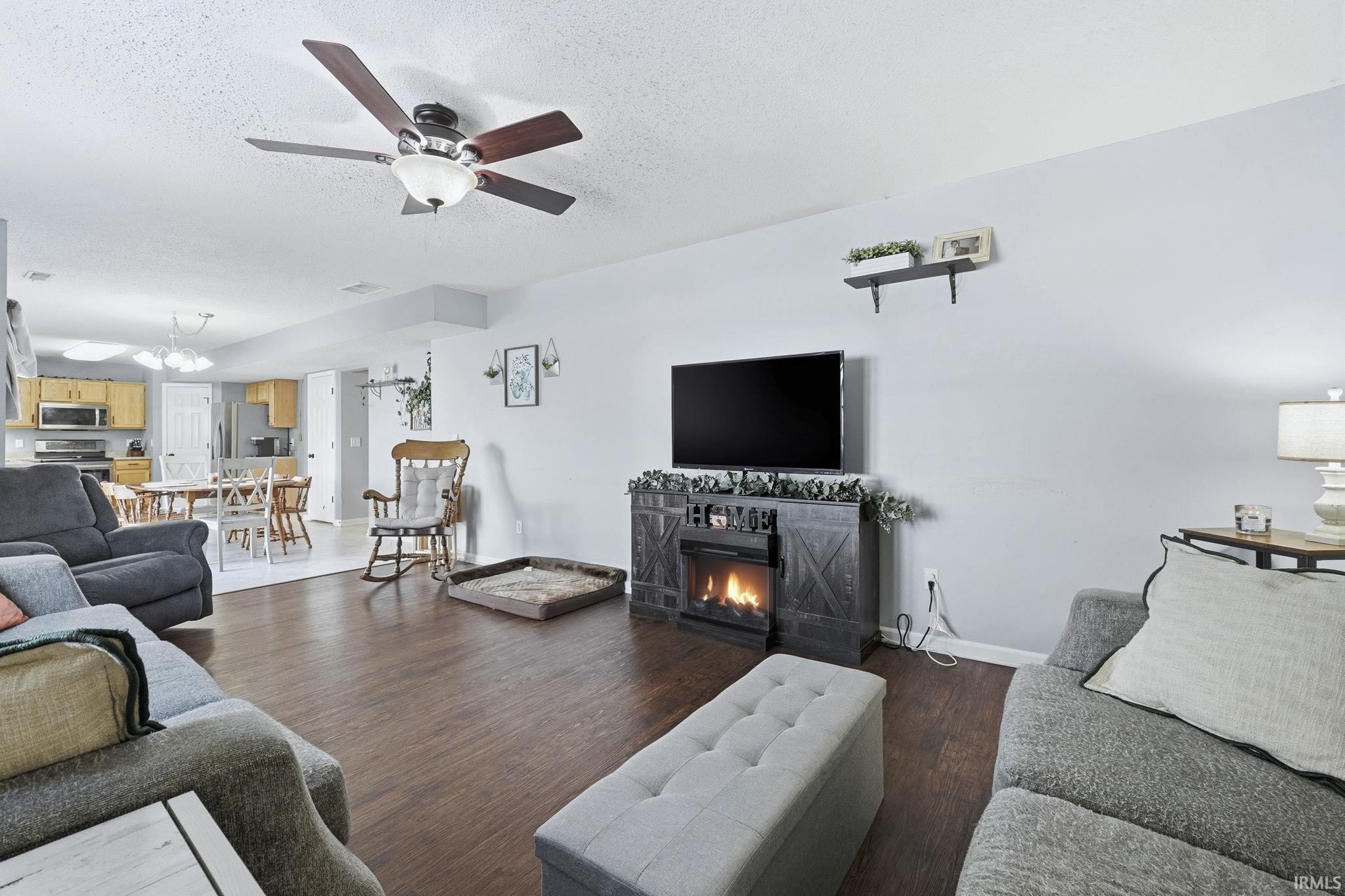 Living room featuring dark wood-type flooring, a ceiling fan, a textured ceiling, and a lit fireplace