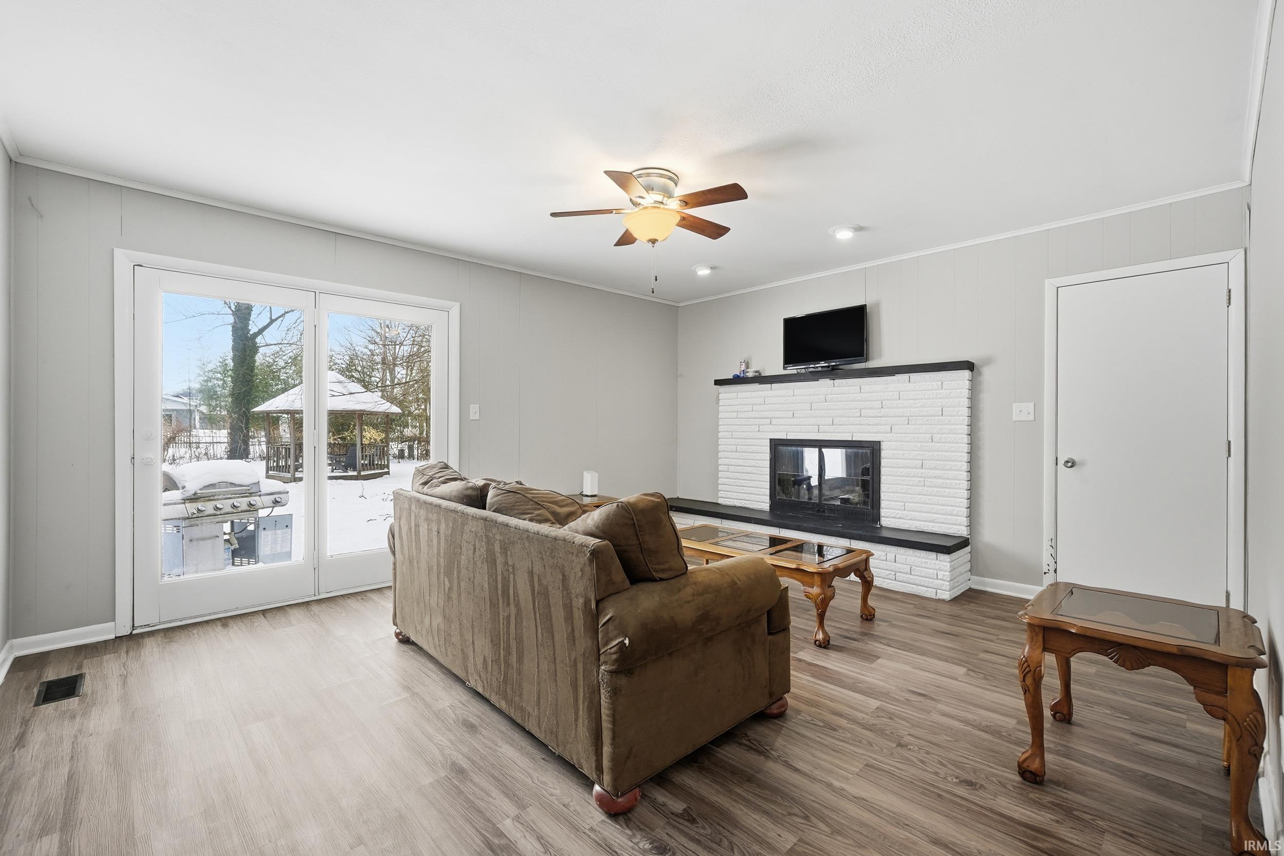 Living room with wood finished floors, a brick fireplace, a ceiling fan, and crown molding