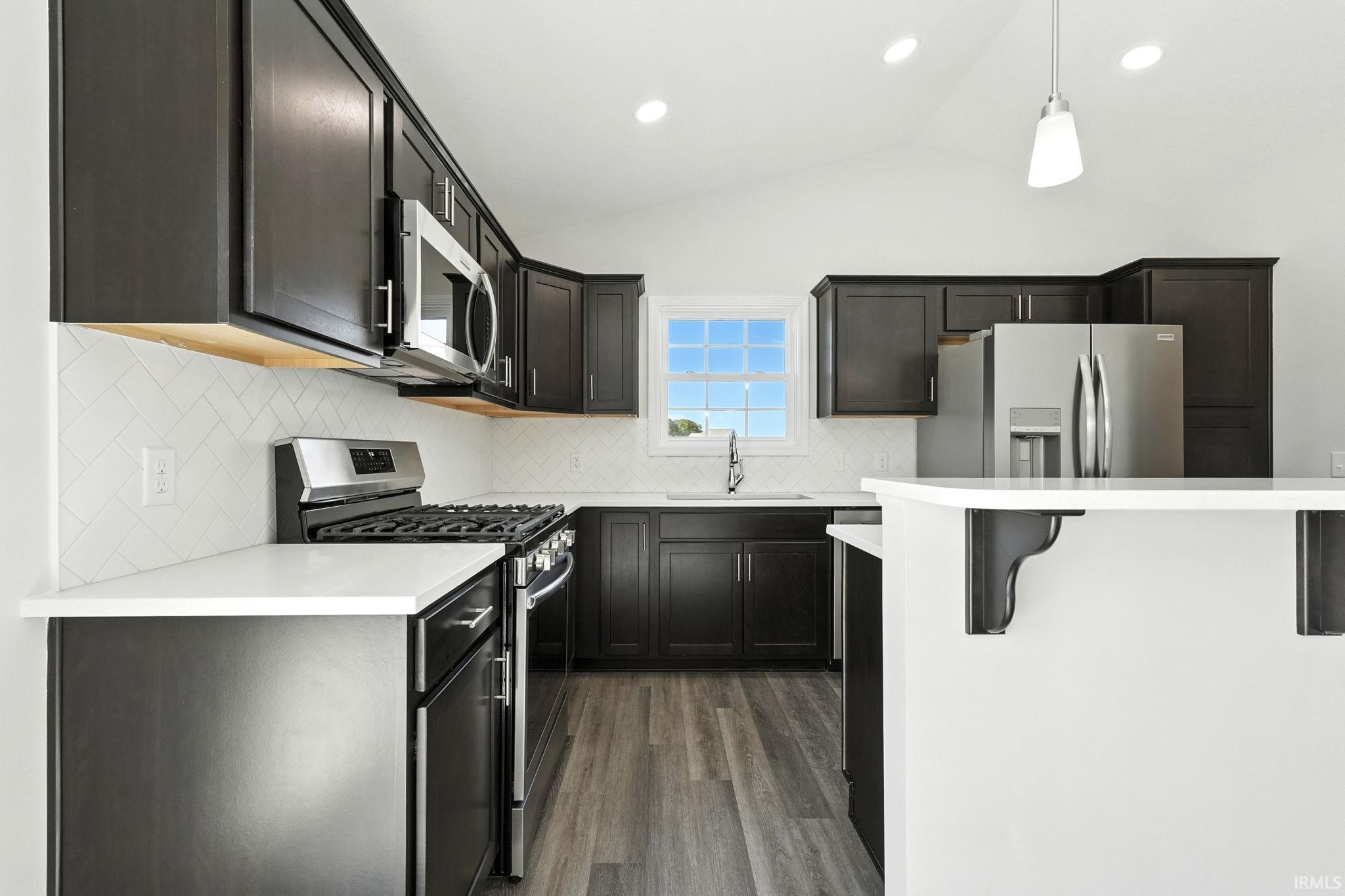 Kitchen with stainless steel appliances, vaulted ceiling, decorative light fixtures, dark wood-style floors, and decorative backsplash