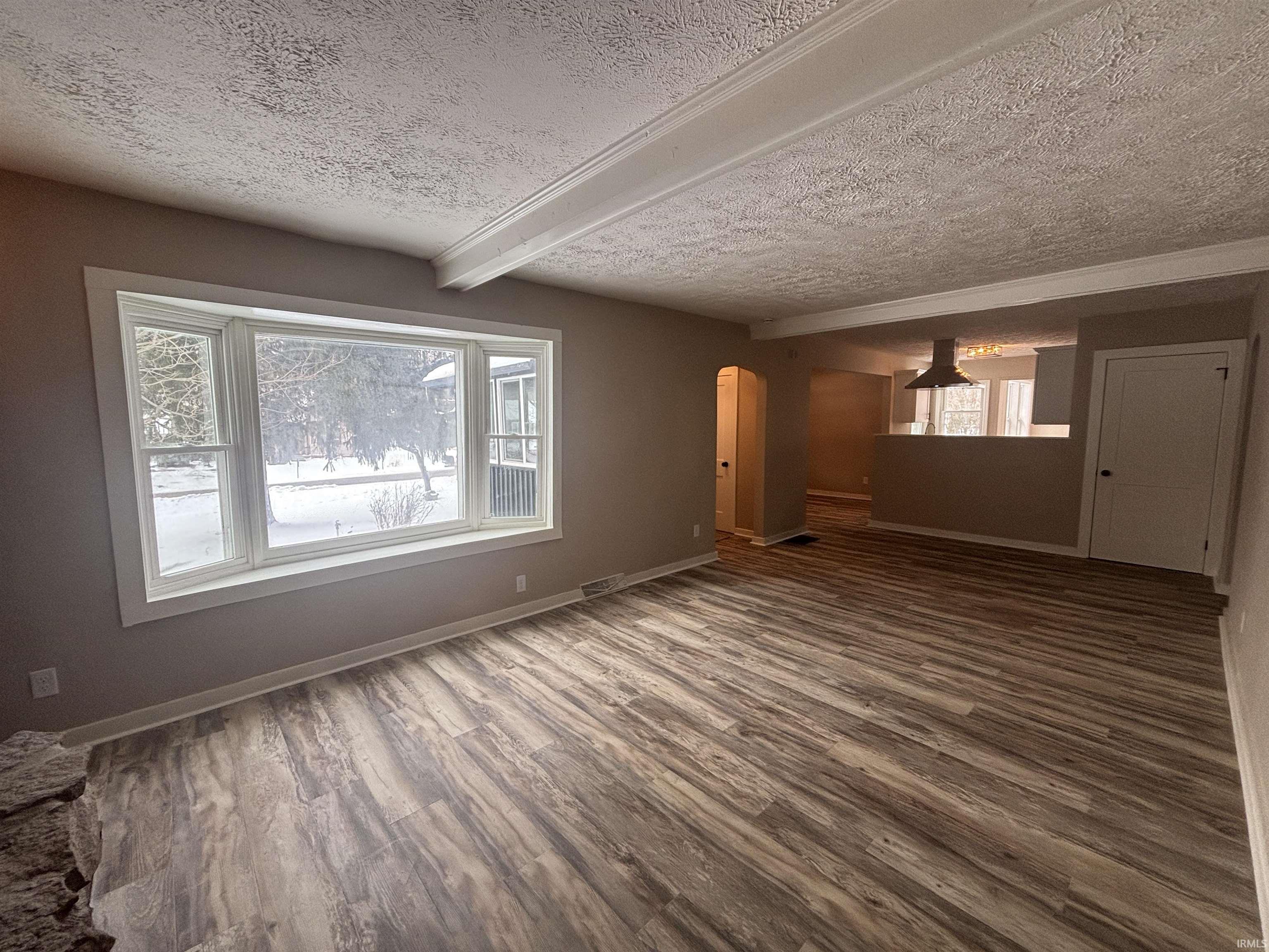 Unfurnished living room with arched walkways, wood finished floors, beamed ceiling, a textured ceiling, and healthy amount of natural light