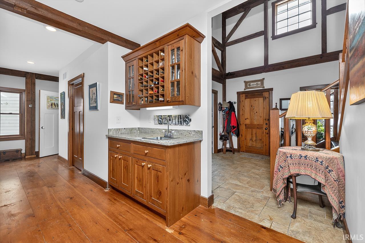 Indoor wet bar featuring glass fronted cabinets, wood finish cabinetry, light stone counters, beamed ceiling, and light wood-style floors