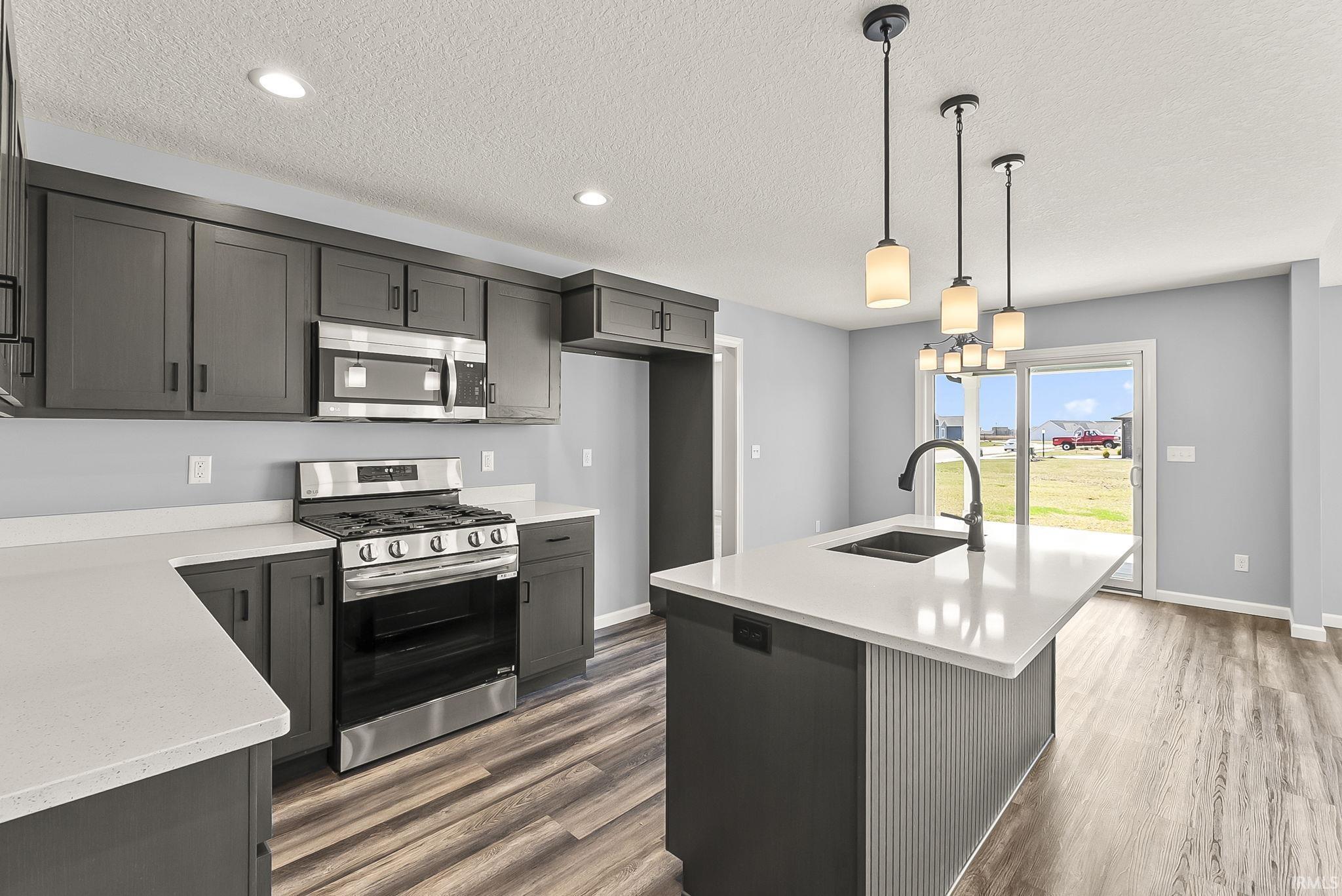 Kitchen with stainless steel appliances, an island with sink, hanging light fixtures, light stone countertops, and a textured ceiling