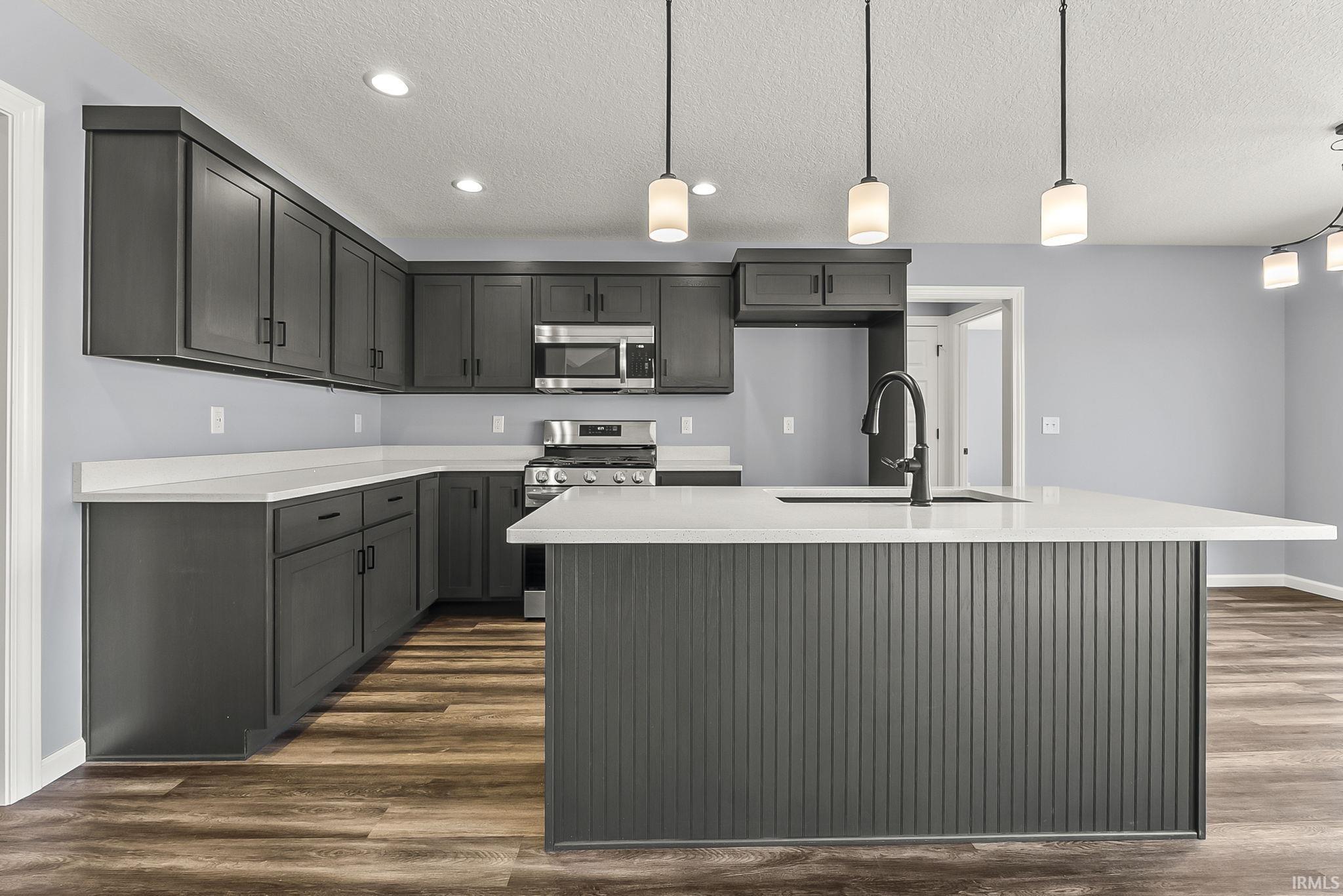 Kitchen featuring a center island with sink, stainless steel appliances, decorative light fixtures, dark wood-type flooring, and a textured ceiling