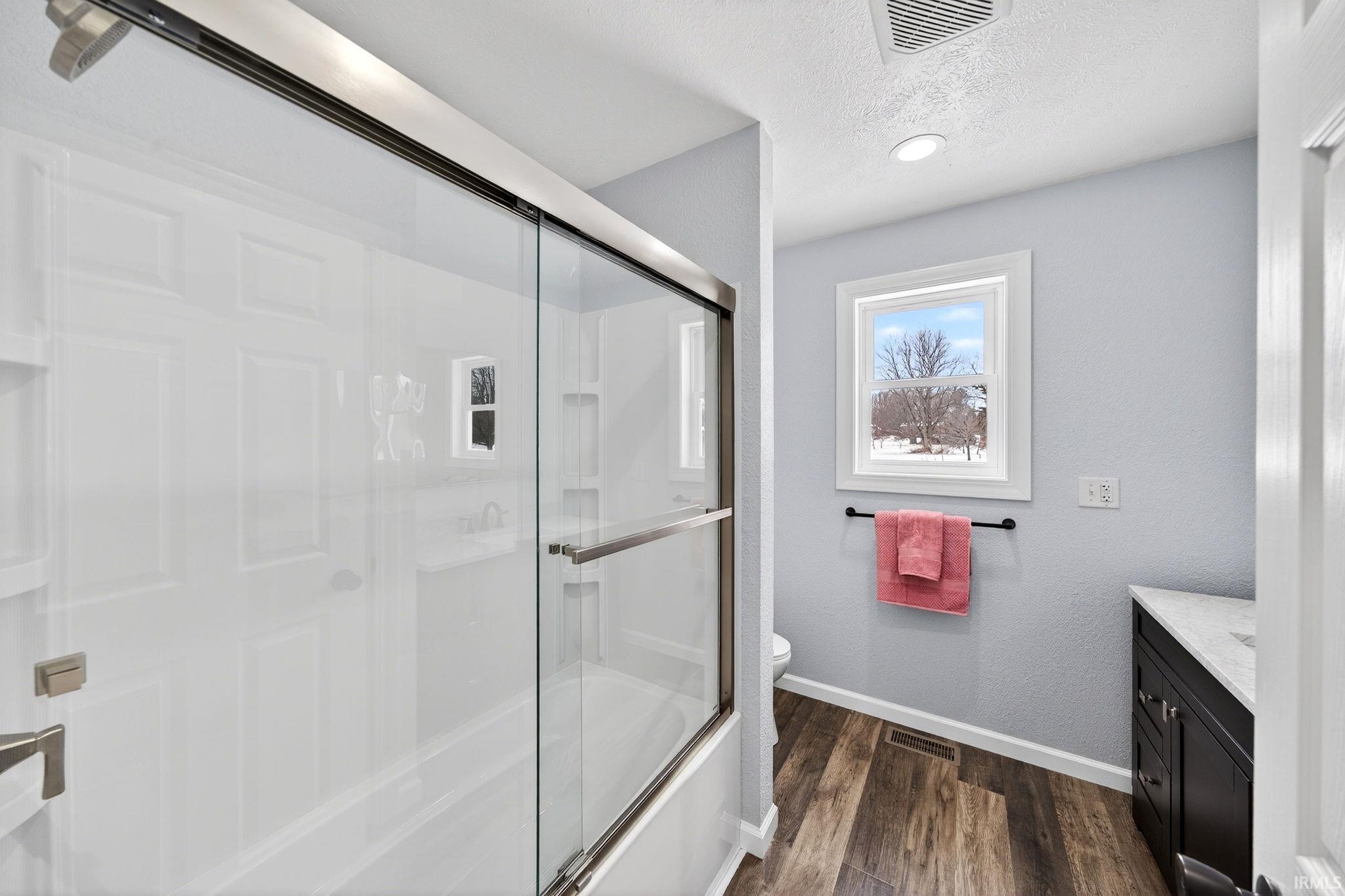 Full bath featuring vanity, a textured ceiling, enclosed tub / shower combo, a textured wall, and dark wood-style flooring