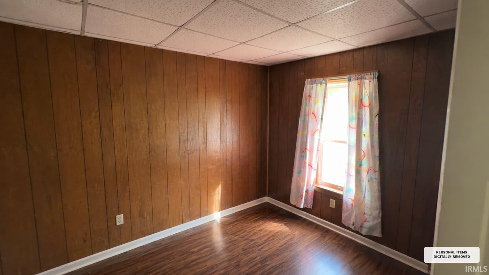 Room featuring full wood-paneling walls, wood-finish flooring, white baseboards, and a grid-pattern ceiling