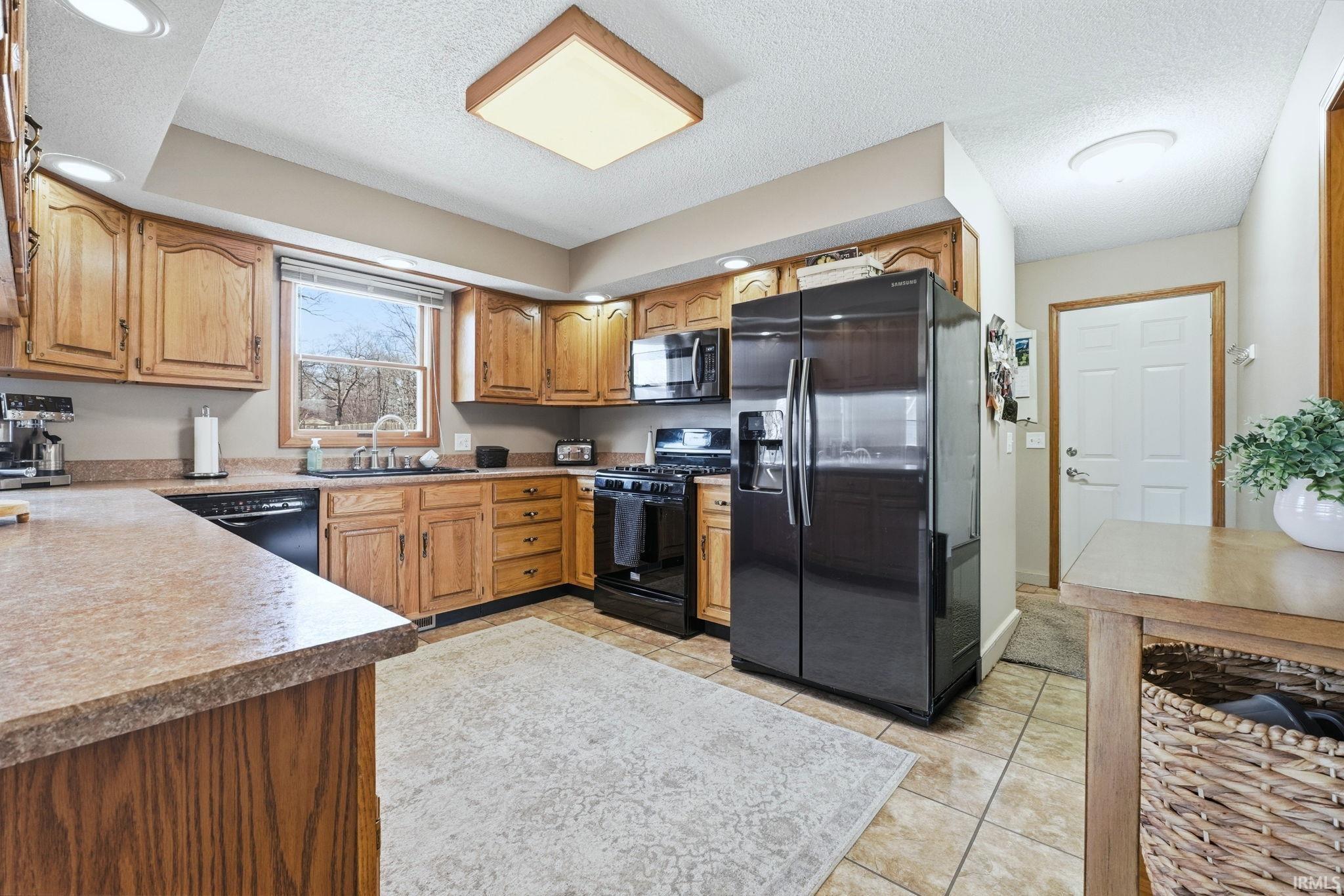 Kitchen with black appliances, a textured ceiling, wood finish cabinets, light tile patterned flooring, and light countertops
