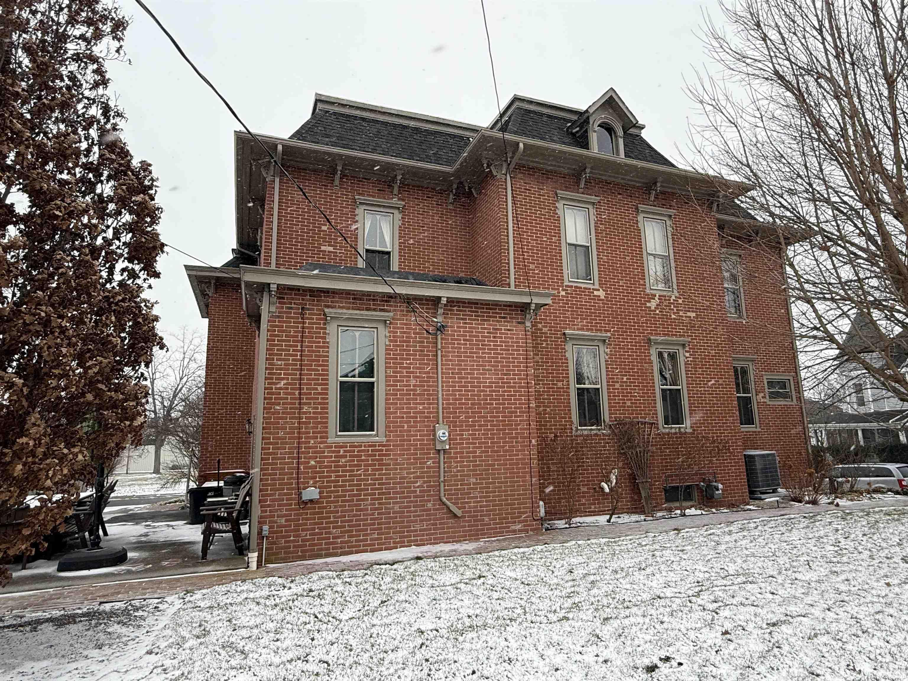 View of snow covered exterior with brick siding, a shingled roof, and mansard roof