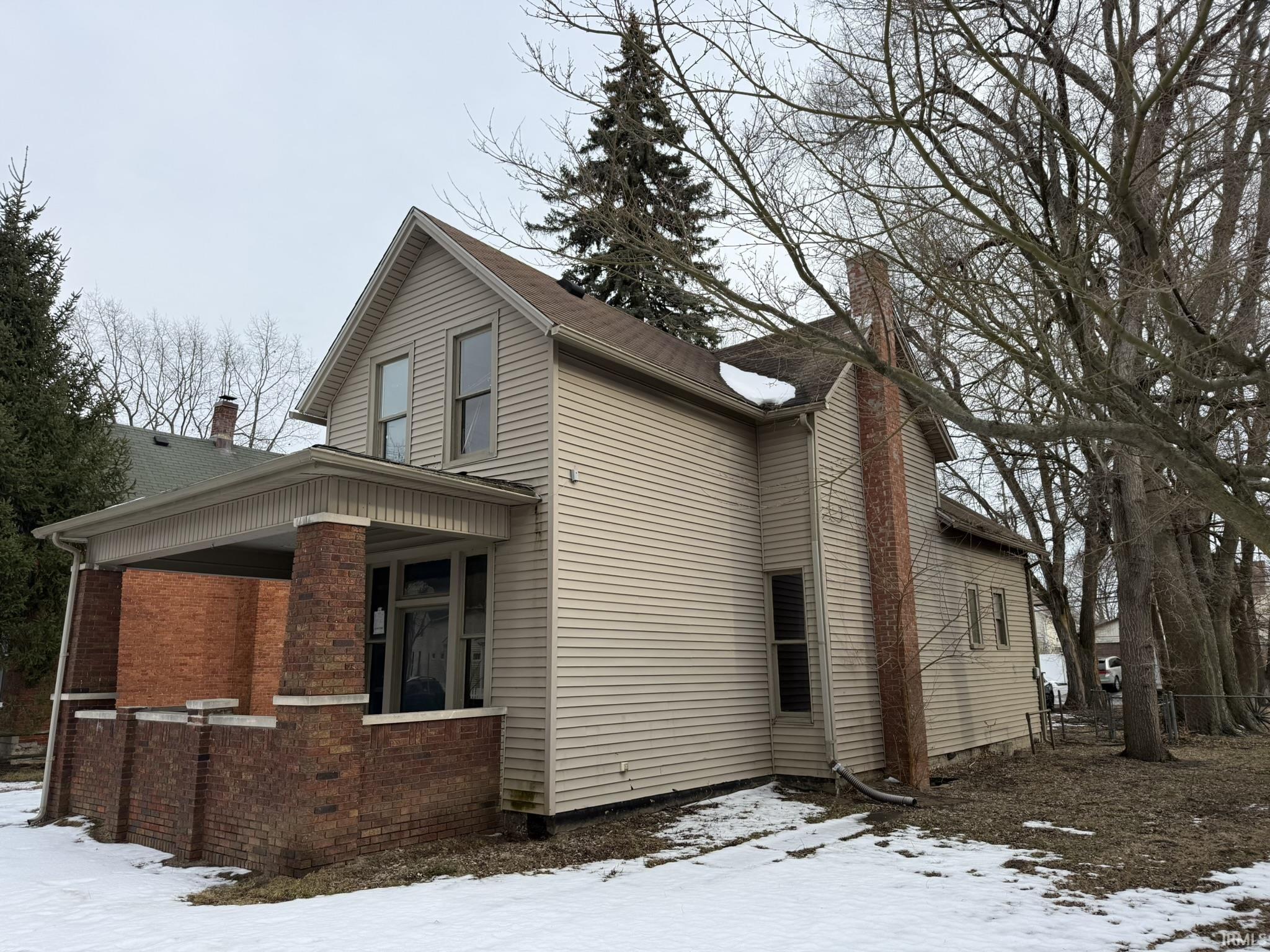 View of snow covered exterior with a chimney, covered porch, and brick siding