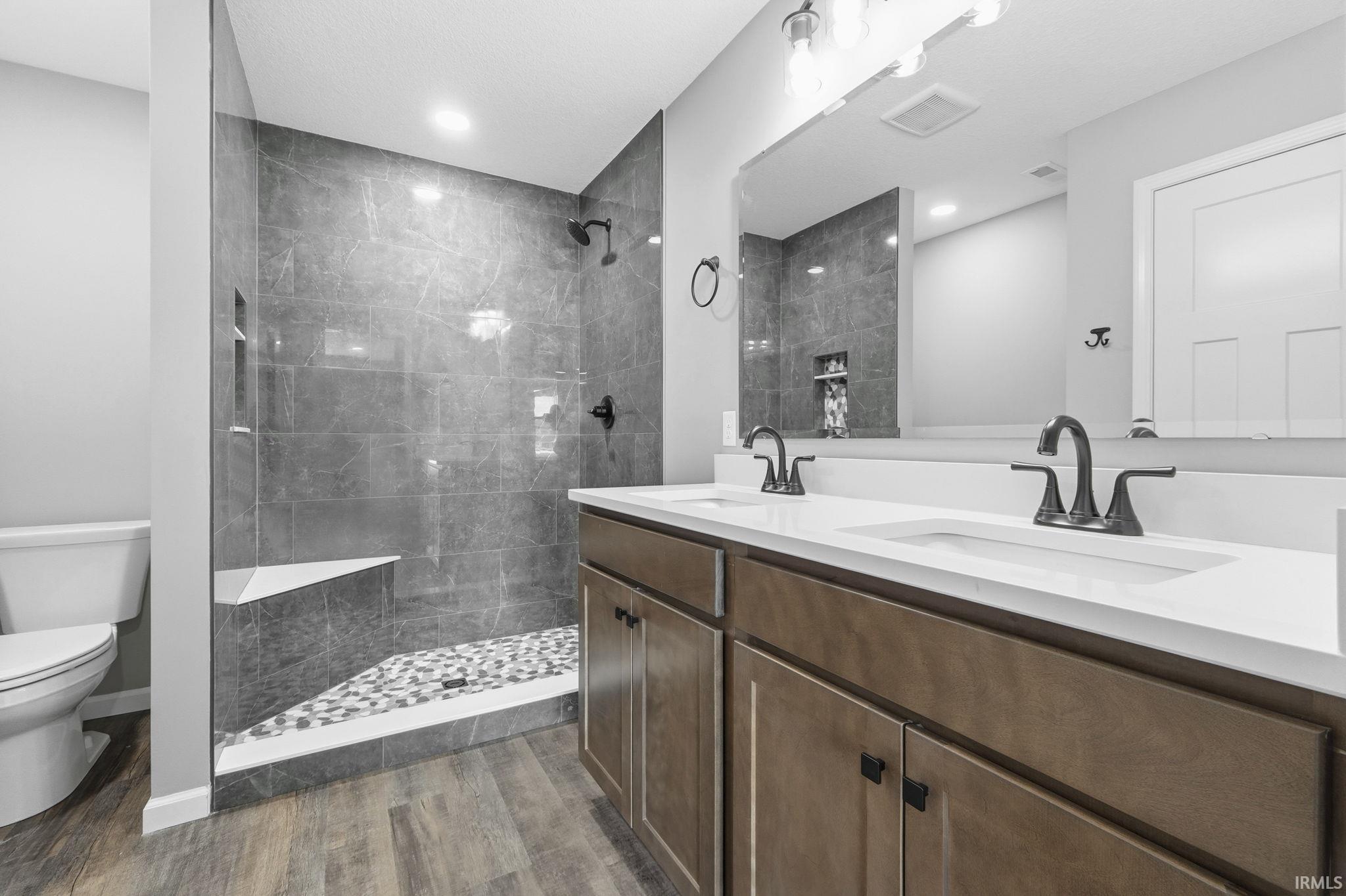 Full bathroom with double vanity, tiled shower, and dark wood-style flooring