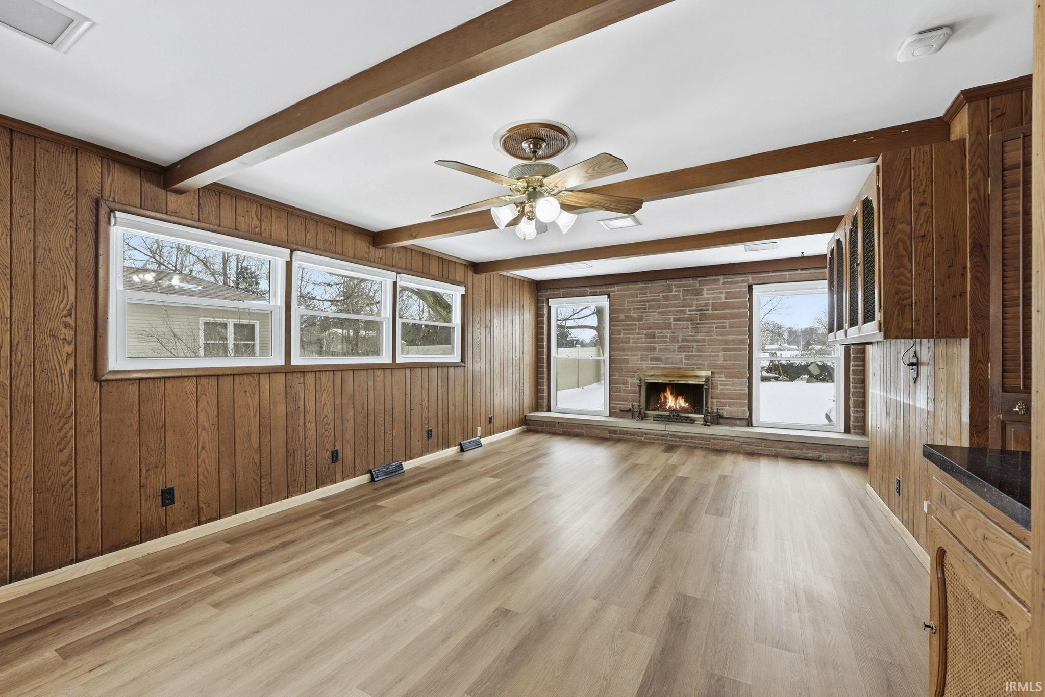 Unfurnished living room featuring wooden walls, beam ceiling, a ceiling fan, a brick fireplace, and light wood-style floors
