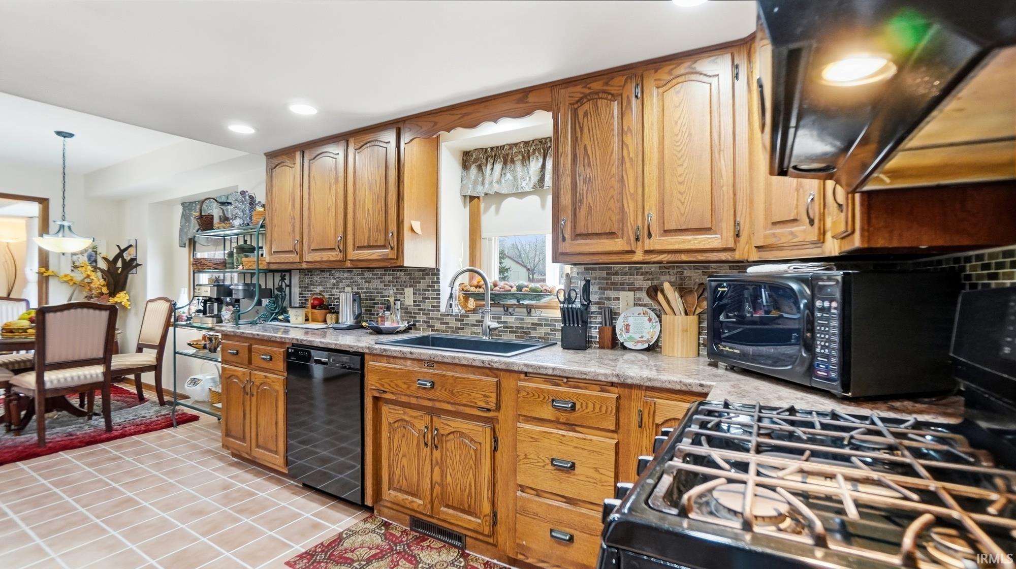 Kitchen featuring wood finish cabinetry, black appliances, range hood, tasteful backsplash, and decorative light fixtures